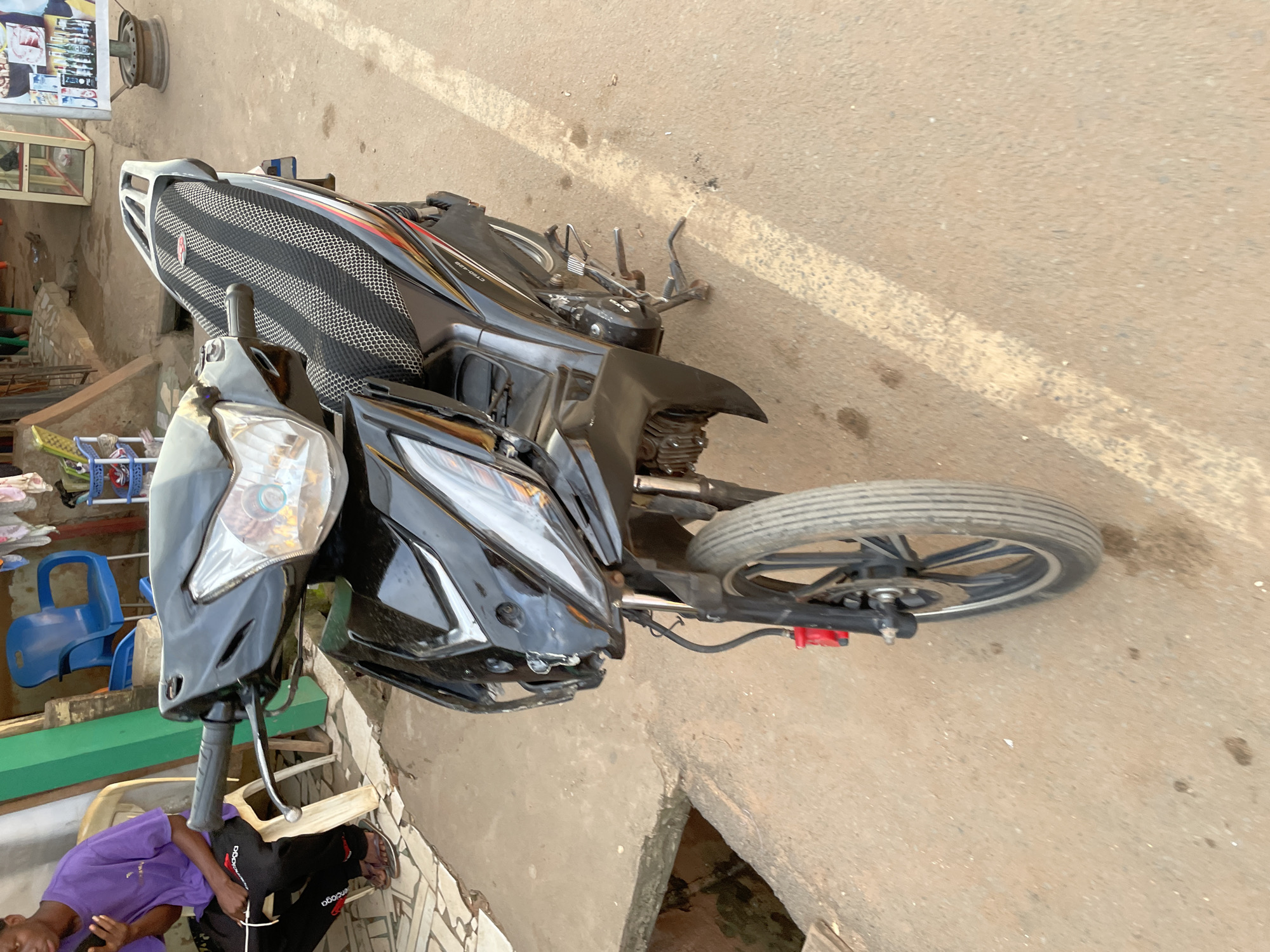 This media file shows a black and red scooter parked on a dirt and concrete surface. The scooter is the primary object, with its front wheel and handlebars visible. Behind the scooter, there are some buildings and what appears to be a market stall with various items displayed. A person wearing a purple shirt and black pants is sitting on a chair to the left of the scooter, partially obscuring the view of the scooter's rear. Another person is visible in the lower left corner of the image. The scene appears to be outdoors, possibly in a public area like a street or market. The lighting suggests it might be daytime, with natural light casting shadows. The environment appears somewhat informal and possibly urban or semi-urban. There is some visible text on the person's pants, which appears to be a brand logo. The overall impression is that of a typical street scene in a Nigerian town or city, with everyday objects and activities.