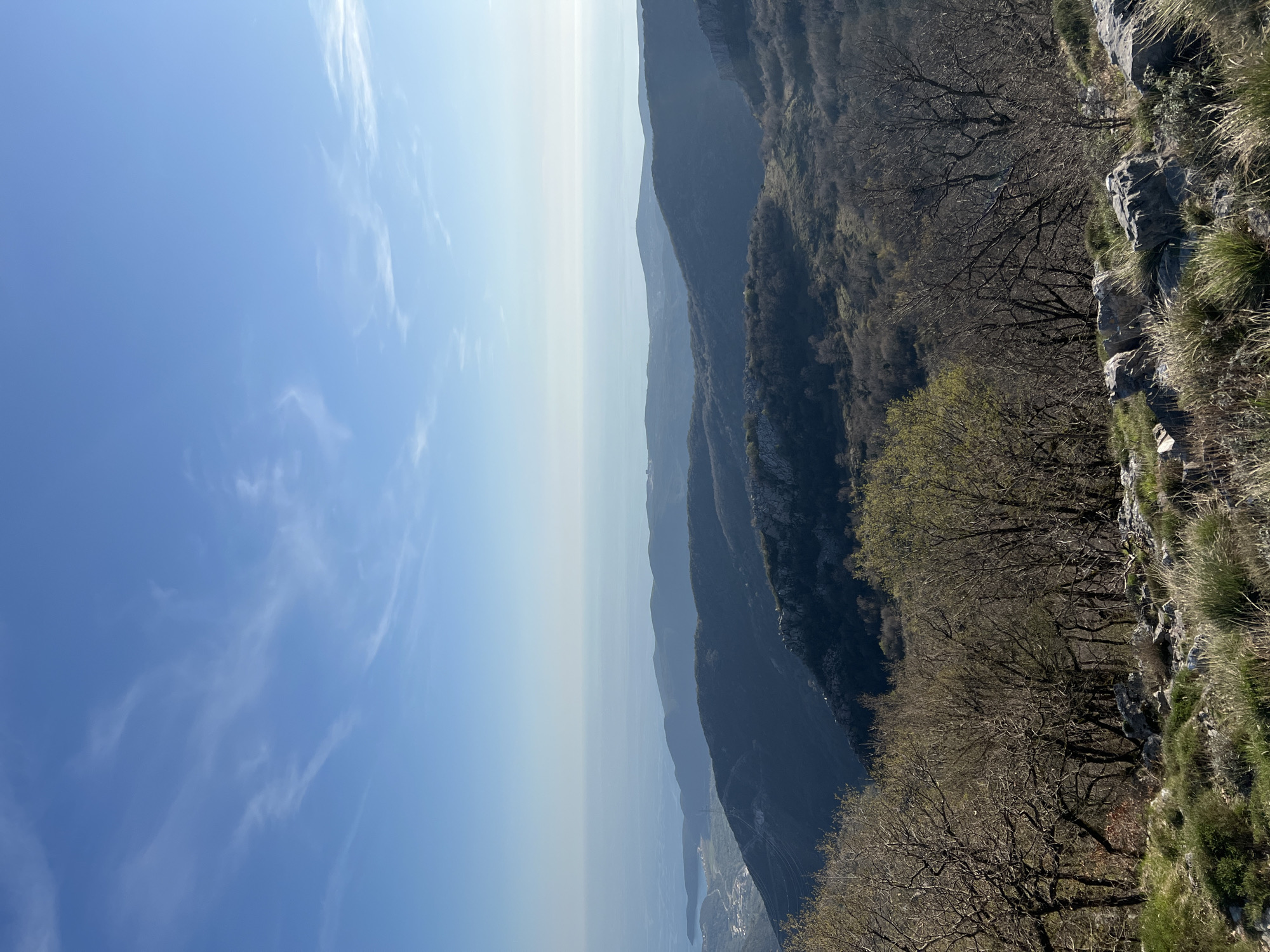 The image depicts a landscape in Dolovi, Montenegro. The view is from a high vantage point, likely a mountainside. The left side of the image shows a blue sky with thin clouds. A body of water stretches horizontally across the middle, obscured by haze, with land visible in the far distance. To the right, mountains descend, covered in varying degrees of vegetation. Bare trees and rocky terrain are visible near the immediate foreground.
