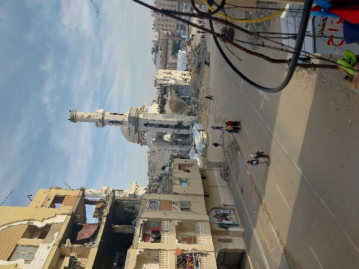 This image depicts a street scene in Khan Yunis, Palestinian Territory, characterized by extensive structural damage under a partly cloudy sky. In the background, a severely damaged mosque stands prominently, its minaret partially intact but its dome and main structure significantly ruined with large sections missing and debris scattered around its base. To the left, a multi-story building exhibits heavy damage, with multiple floors exposed, walls blown out, and structural elements twisted. Laundry hangs from a balcony railing on a lower, less damaged section of this building. The foreground features a paved road with white lane markings. Several individuals and small groups are present on the street and surrounding areas: two people walk in the left mid-ground, a group of approximately five individuals stands in the middle of the road further back, and additional solitary figures traverse areas of rubble in the background. Piles of what appear to be corrugated metal sheets are visible amidst the debris behind the mosque. In the immediate foreground, rusty metal bars and cables, possibly power lines, hang, with colorful clothes clips attached. A partially visible sign with Arabic text is located below these cables.