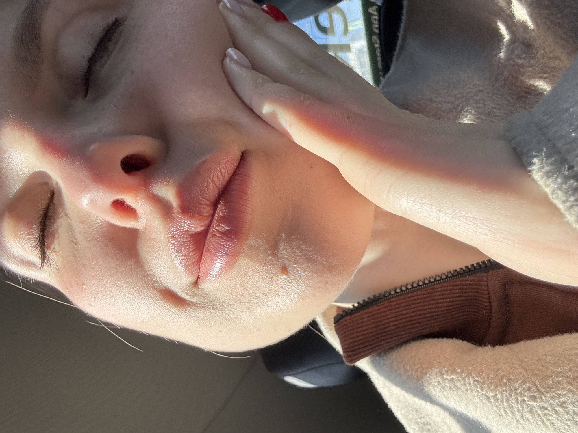 A close-up, low-angle shot of a fair-skinned person's face with closed eyes. Part of a hand is touching the cheek. The lips are full and appear to have lip gloss or lipstick applied. The person is wearing a cream-colored, fuzzy sweater with a brown collar visible near the neck. Red nail polish is visible on one finger. The location is unknown, as there are no identifying details, so the location in Moscow, Russia, cannot be confirmed.