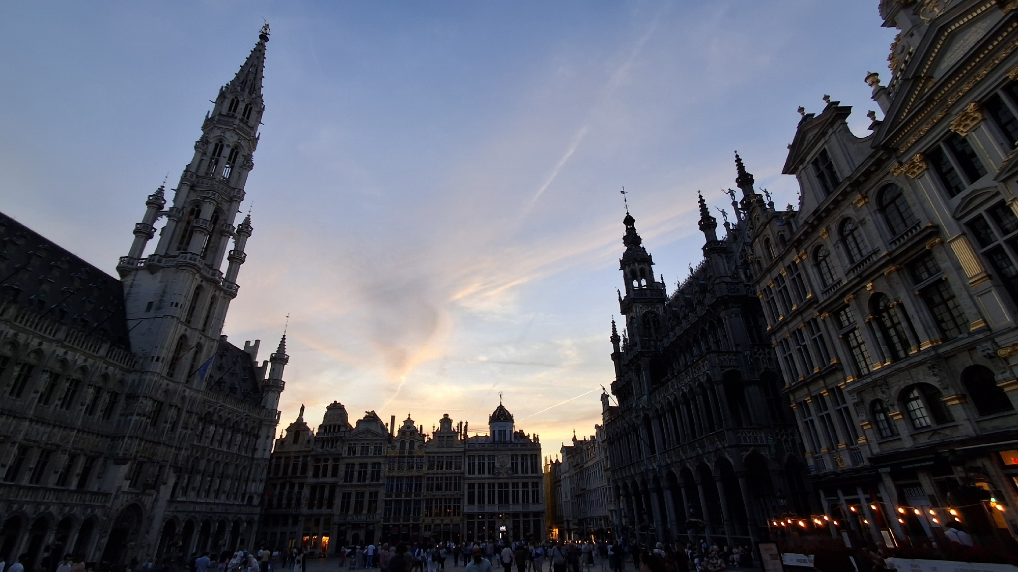 The image shows a wide-angle view of a European town square at dusk. The sky is a pale blue with wispy clouds tinged with orange and pink, suggesting the sun has recently set. The architecture is ornate and gothic, with tall spires and intricately decorated buildings lining the square. In the foreground, a crowd of people is visible, casually strolling and gathering. Numerous outdoor lights, strung along the fronts of buildings, illuminate the scene. The overall impression is one of a lively public space at the end of the day.