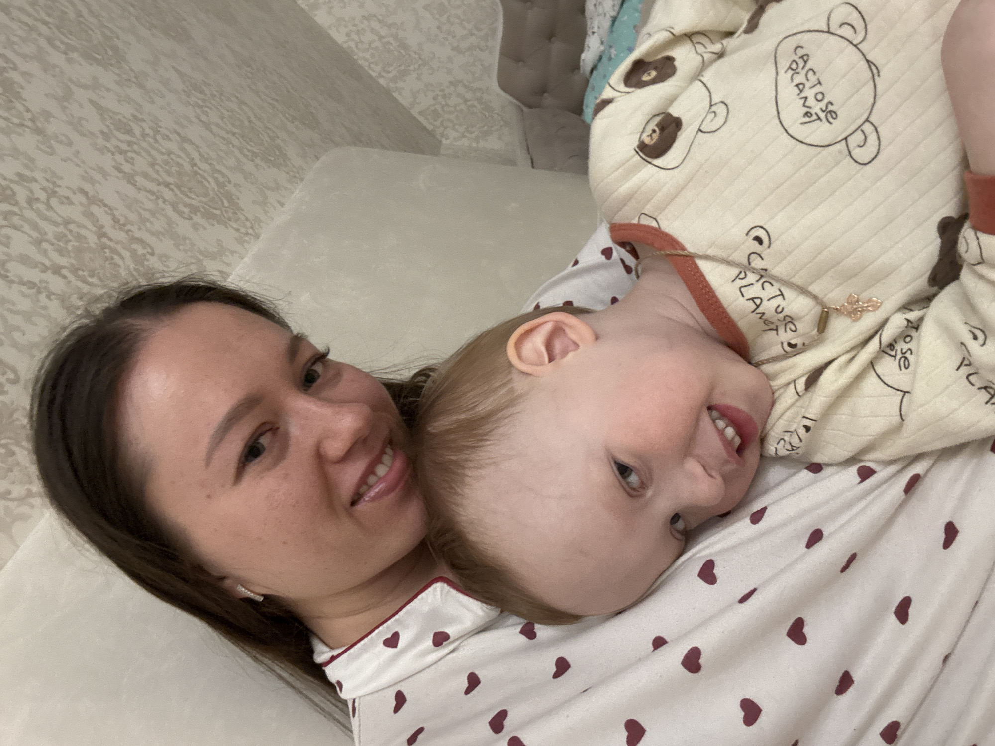 A woman and a young child are lying down, possibly on a bed or soft surface. The woman has dark hair and is smiling. The child is wearing a light-colored pajama top with brown heart patterns, and an off-white top with brown bear illustrations. The bears are labeled "Cactose Planet". The child has light hair and is also smiling. The child wears a necklace, a gold-colored chain with a clover or flower shaped pendant. The background includes a textured wall and what appears to be a cushioned headboard or piece of furniture. The location is Ферма, Russia.