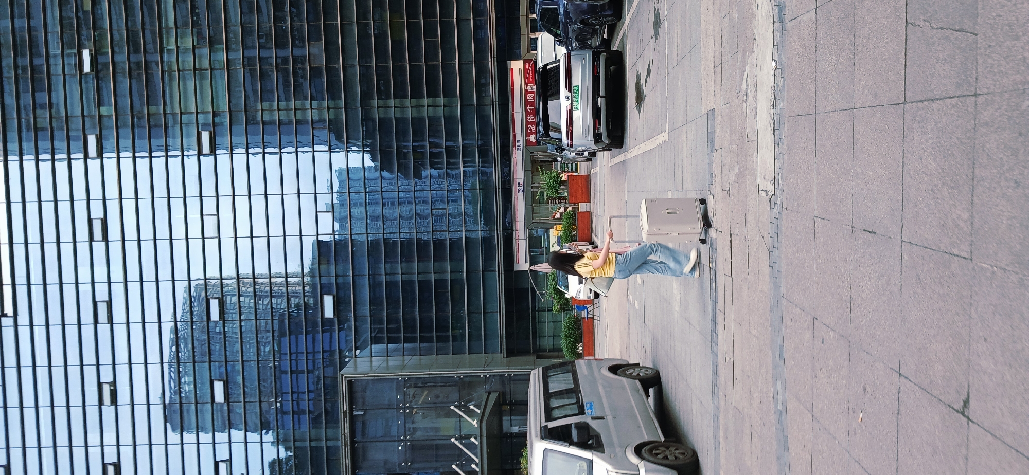 A full shot depicts a young woman pulling a white suitcase along a sidewalk in Jiulongpo District, China. The woman is wearing a yellow t-shirt and blue jeans. She is walking from right to left.
To the woman’s left is a modern building with a mirrored glass facade reflecting an adjacent building. Parallel to the walkway, cars are parked along the curb. A silver van is parked closer to the camera's perspective. The sidewalk is paved with gray, square paving stones.