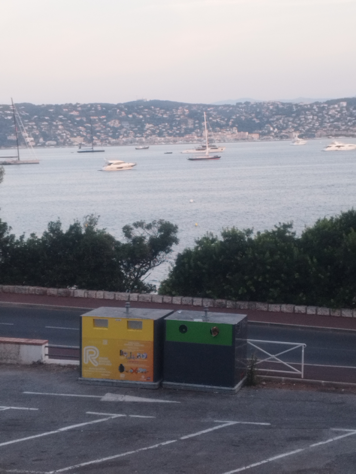 The image captures a scene in Antibes, France, overlooking a bay dotted with various boats and yachts. In the foreground, two recycling bins stand side-by-side on asphalt paving marked with white lines. The left bin is predominantly yellow with orange accents and features a prominent "R" logo. The right bin is dark grey with a green top section. A low stone wall runs along the edge of the road behind the bins, separating it from dense green foliage. Beyond the foliage, a road is visible, and further in the distance, the coastline of Antibes is lined with buildings, nestled against a backdrop of green hills. The sky above is overcast.