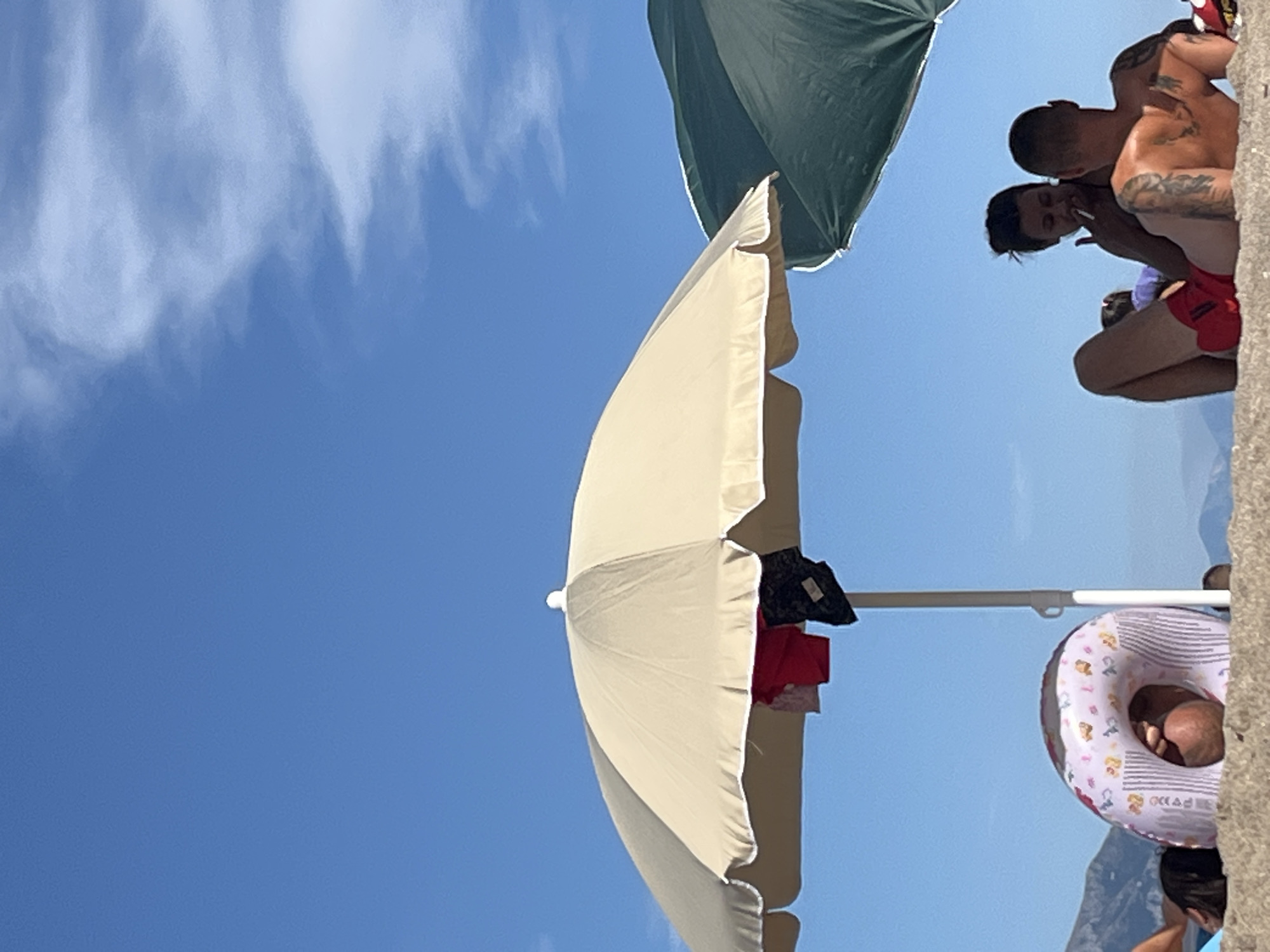 In Prijevor, Montenegro, on a sandy beach under a bright blue sky with wispy clouds, a scene of relaxation unfolds. Two beach umbrellas, one a light tan and the other dark green, offer shade. Clothing, including red and patterned shorts and a black garment, is draped over the pole of the tan umbrella. In the foreground, partially obscured by a brightly colored inflatable ring with a cartoon pattern, a person's head and shoulders are visible, resting on the sand. Behind the inflatable ring, a man and a woman are seated together under the green umbrella. The man, facing away from the camera, has tattoos on his back. The woman, facing the man, appears to be smoking and looking down. They are both dressed in swimwear. The background shows the hazy outline of mountains against the horizon.