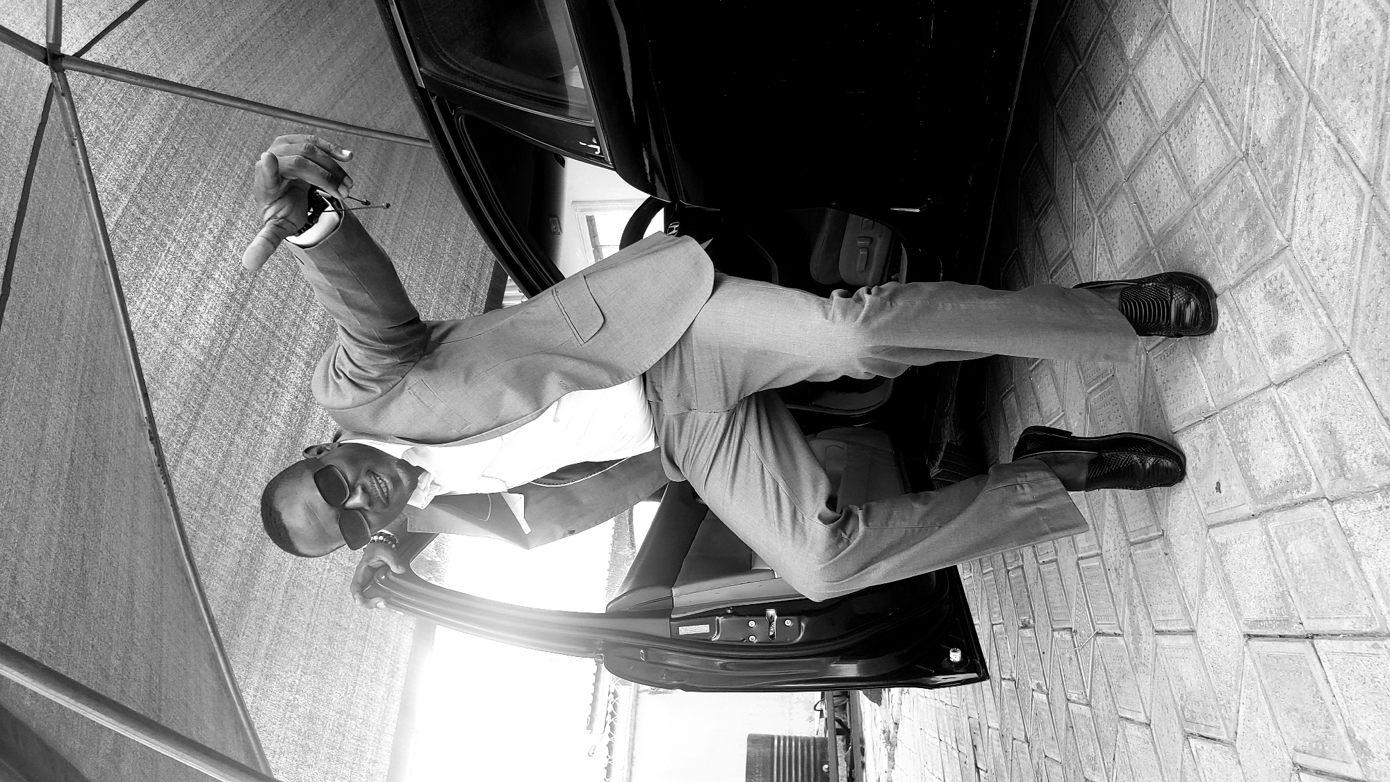 This black and white photograph captures a stylishly dressed man posing next to an open black car during the daytime in Sabon Tasha, Nigeria. The man, with dark skin, is wearing a light-colored suit comprising a jacket and trousers over a white shirt, complemented by black sunglasses and black formal shoes. He is smiling broadly, conveying a sense of confidence or happiness. His body is angled towards the viewer, leaning against the open car door. His left leg is firmly on the paved ground, while his right leg is bent and partially inside the car's interior. His right arm is raised, holding what appears to be car keys or another pair of glasses, and his left arm rests casually on the car's door frame.
The setting is an outdoor paved area with light-colored, patterned tiles on the ground. Above, a tent-like structure with a mesh fabric provides shade, suggesting a carport or a canopy. The bright lighting indicates a clear day. No discernible text is visible in the image.
