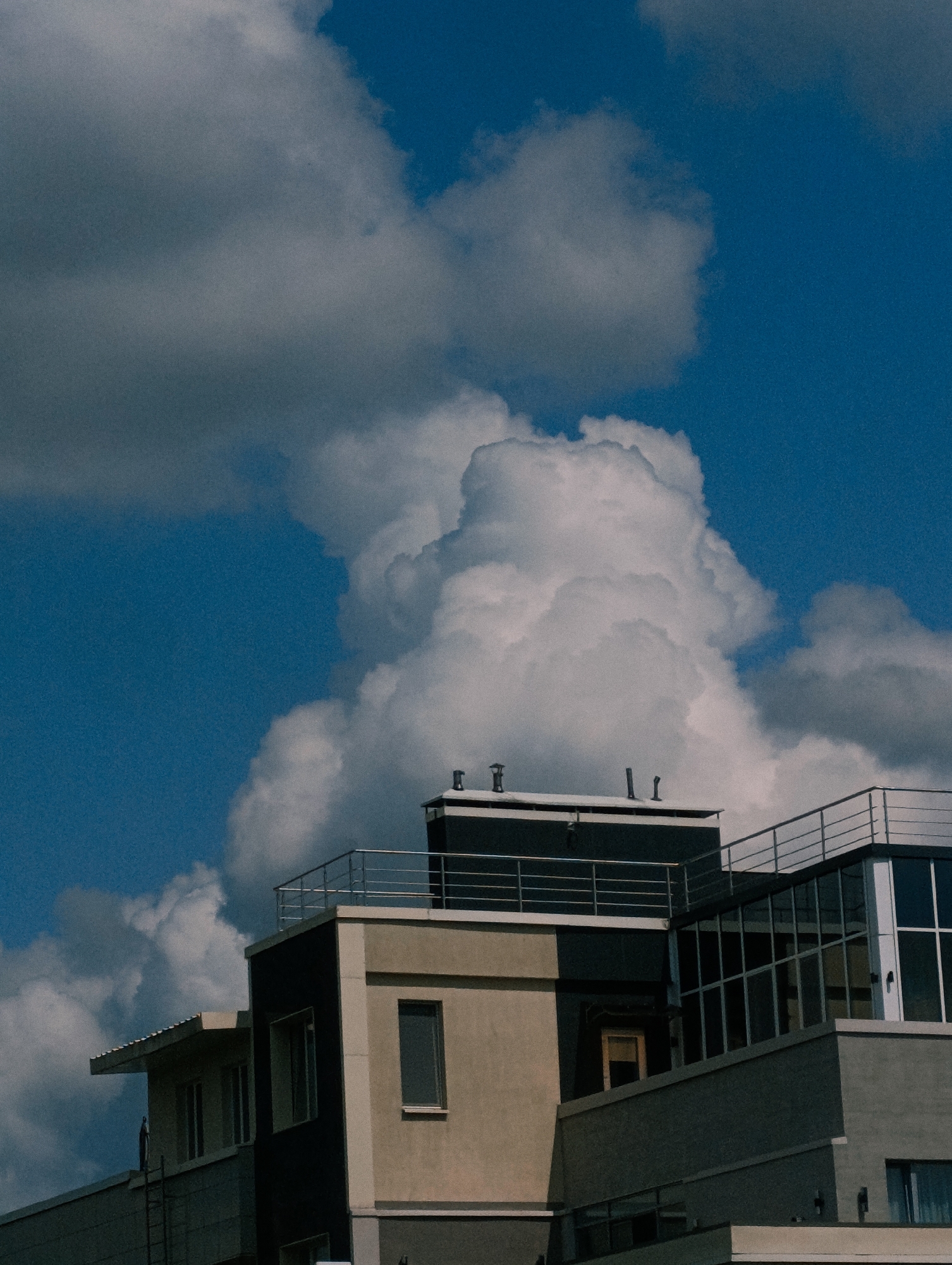 An outdoor, medium shot captures the upper floors of several modern buildings against a bright blue sky filled with large, fluffy white clouds and some darker gray clouds. The buildings have a contemporary architectural style, featuring flat roofs, balconies with railings, and large windows. One building section is a light tan color, another is dark gray, and a section with large glass panels reflects the sky. There are several ventilation pipes or chimneys visible on the roof of a dark section. A ladder is partially visible on the side of one building. The scene is static with no people or discernible actions. The location is identified as Unknown, Unknown.