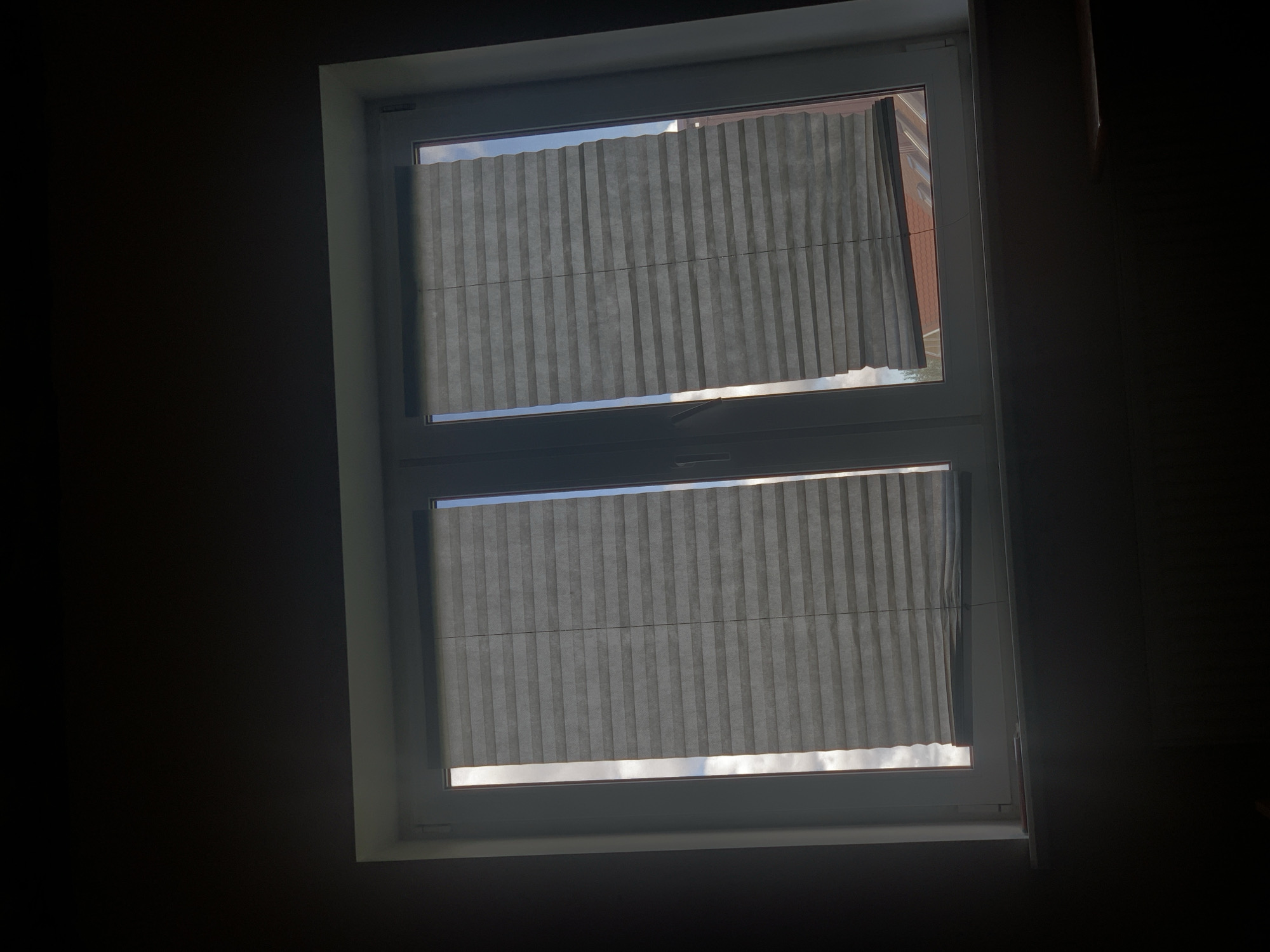 The image shows a closed window with pleated blinds. The window is divided into two sections, each covered by a blind. Through the blinds, the sky and some rooftops are visible. The interior of the room is dark, suggesting it is either nighttime or the blinds are drawn to block out light. There are no people or actions visible in the image. The location is identified as Minsk, Belarus.