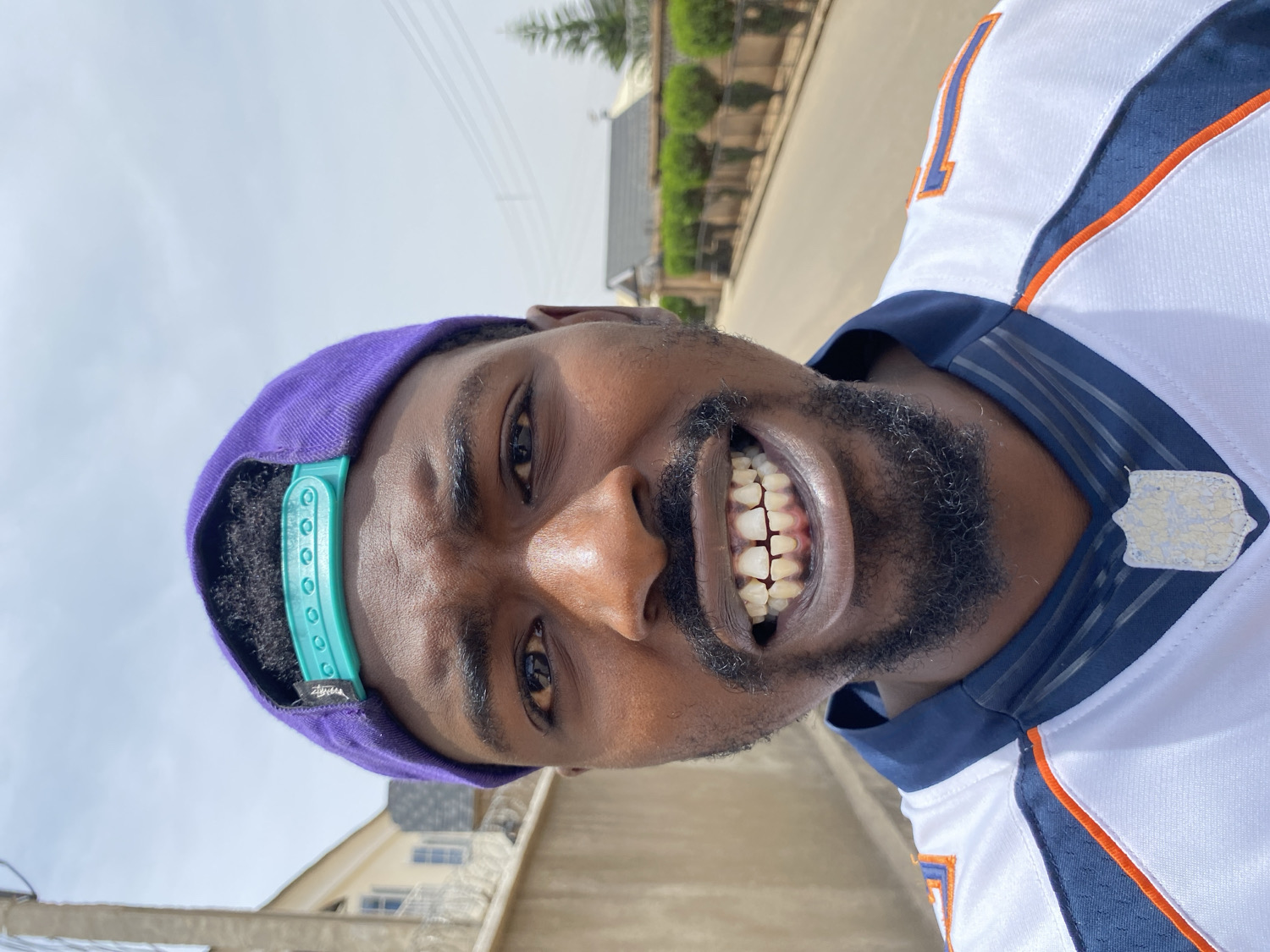 A close-up, portrait-oriented photograph (rotated 90 degrees counter-clockwise) captures a smiling Black man outdoors in Jos, Nigeria, under bright daylight. He is looking directly at the camera with a wide, joyful grin, showcasing his teeth. The man has a full beard and short dark hair, partially covered by a purple baseball cap worn backward. The cap features a light teal/turquoise plastic snapback adjuster with the brand name "STüSSY" visible on a small tag. He is wearing a white sports jersey, likely an American football jersey, with navy blue and orange stripes and accents; the number "1" is visible on the upper right arm/shoulder area. A worn, shield-shaped patch or emblem is located on the jersey's collar. The background shows a bright, clear or lightly cloudy sky, with indistinct buildings and hints of green foliage, suggesting an urban or suburban setting. The man's expression conveys happiness and friendliness.