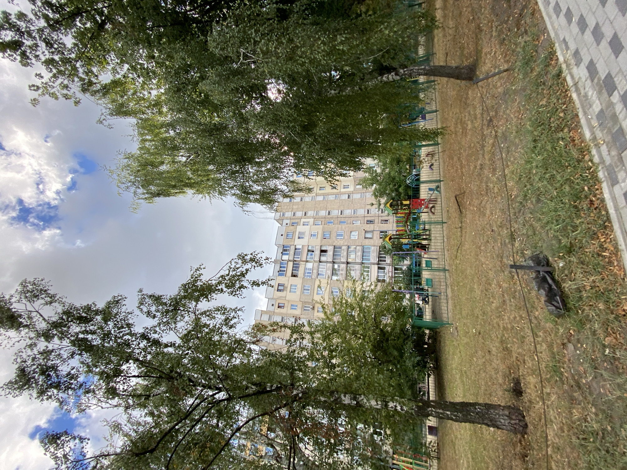 The image captures a view of a residential building and a playground on an overcast day. In the foreground, dry grass and fallen leaves cover the ground. A dark, trash-like object is visible on the ground, partially obscured by a wire fence. The fence encloses a playground area with colorful structures, including swings and slides. Beyond the playground, a multi-story apartment building stands tall, its facade featuring numerous windows and balconies. Several trees with green foliage are scattered throughout the scene, with one prominently positioned in the left foreground. The sky above is filled with clouds, with patches of blue sky peeking through.