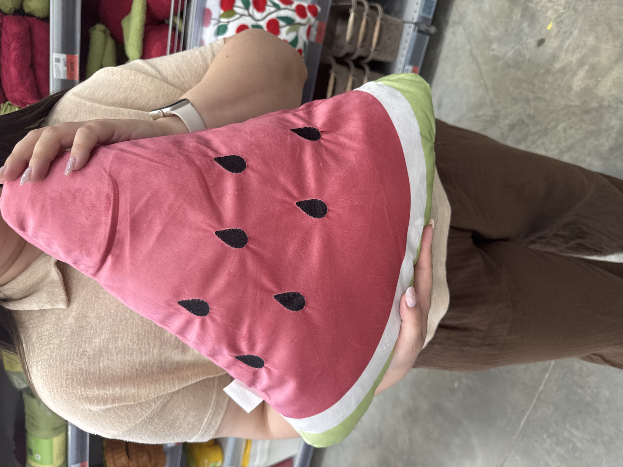 A person is holding a plush pillow shaped like a slice of watermelon. The pillow is primarily pink with black seeds and green and white rind. The person is wearing a light-colored, textured long-sleeved shirt and brown pants. A white smartwatch is visible on their left wrist. The background shows a retail environment with shelves displaying various items, including what appear to be towels or textiles in shades of pink and green. The floor is a light gray, concrete-like surface. The lighting suggests an indoor setting, likely a store, during daytime. The image is taken from a high-angle perspective, looking down at the person and the pillow. There is no visible text in the frame. The overall impression is that of someone shopping or browsing in a store, holding a decorative item.