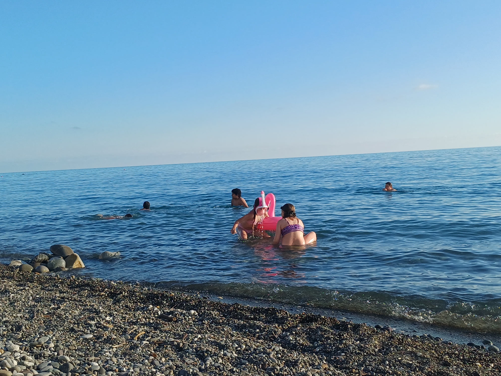 A medium shot captures a serene beach scene in Sochi, Russia, under a clear blue sky. The foreground is dominated by a pebble-strewn beach, with a gentle wave lapping at the shore. In the tranquil blue water, several individuals are enjoying a summer day. Two children, a boy and a girl, are seated on a pink inflatable flamingo, with the girl wearing a snorkel mask. Nearby, another child is swimming in the sea, while a fourth person is visible further out in the water. The overall impression is one of peaceful recreation on a warm, sunny day by the Black Sea.