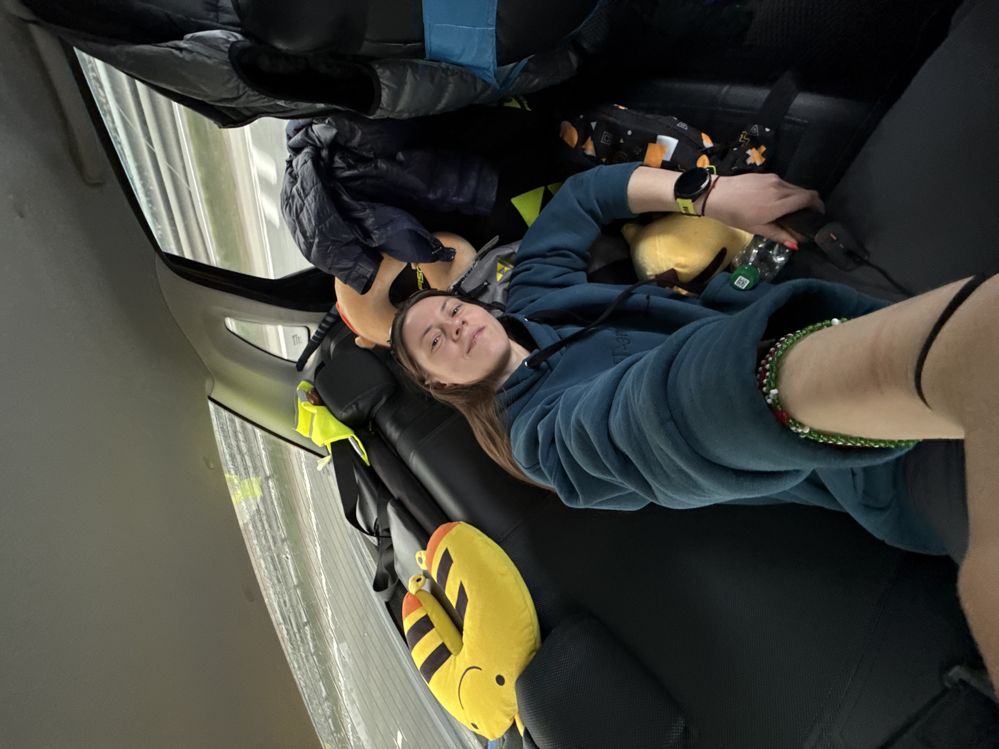 A young woman is depicted lying down in the rear interior of a vehicle, likely a car, with the camera angled to capture her and the surrounding space. She wears a teal blue hoodie and has medium-length brown hair, looking directly towards the viewer with a neutral expression.
The car's interior features dark seats and grey trim. Through the window on the left, a blurred outdoor scene with light-colored lines suggests the vehicle is in motion, possibly on a highway or road. Above the woman, various items are stored, including a dark puffer jacket, a backpack with blue accents, and a geometrically patterned bag with orange, black, white, and yellow designs.
Around the woman, comfort items are visible. A yellow, toucan-shaped neck pillow with black and orange stripes and a smiling face rests beside her. Another yellow plush toy and a small, light-colored plush head are also present. On her left wrist, she wears a smartwatch with a yellow band. Her left hand is near a small green bottle and a dark electronic device, possibly a power bank, connected by a black cable. A colorful beaded bracelet (green, red, white) is visible on her right ankle.
The scene suggests the woman is resting or traveling in the back of the car. The visible motion blur from outside the window reinforces the idea of a journey. The location context provided is Покровское, Russia. No significant legible text is clearly visible on any items, though a faint "FISCH" might be present on a backpack and "TH" or "TR" on a car panel. The lighting indicates it is daytime.