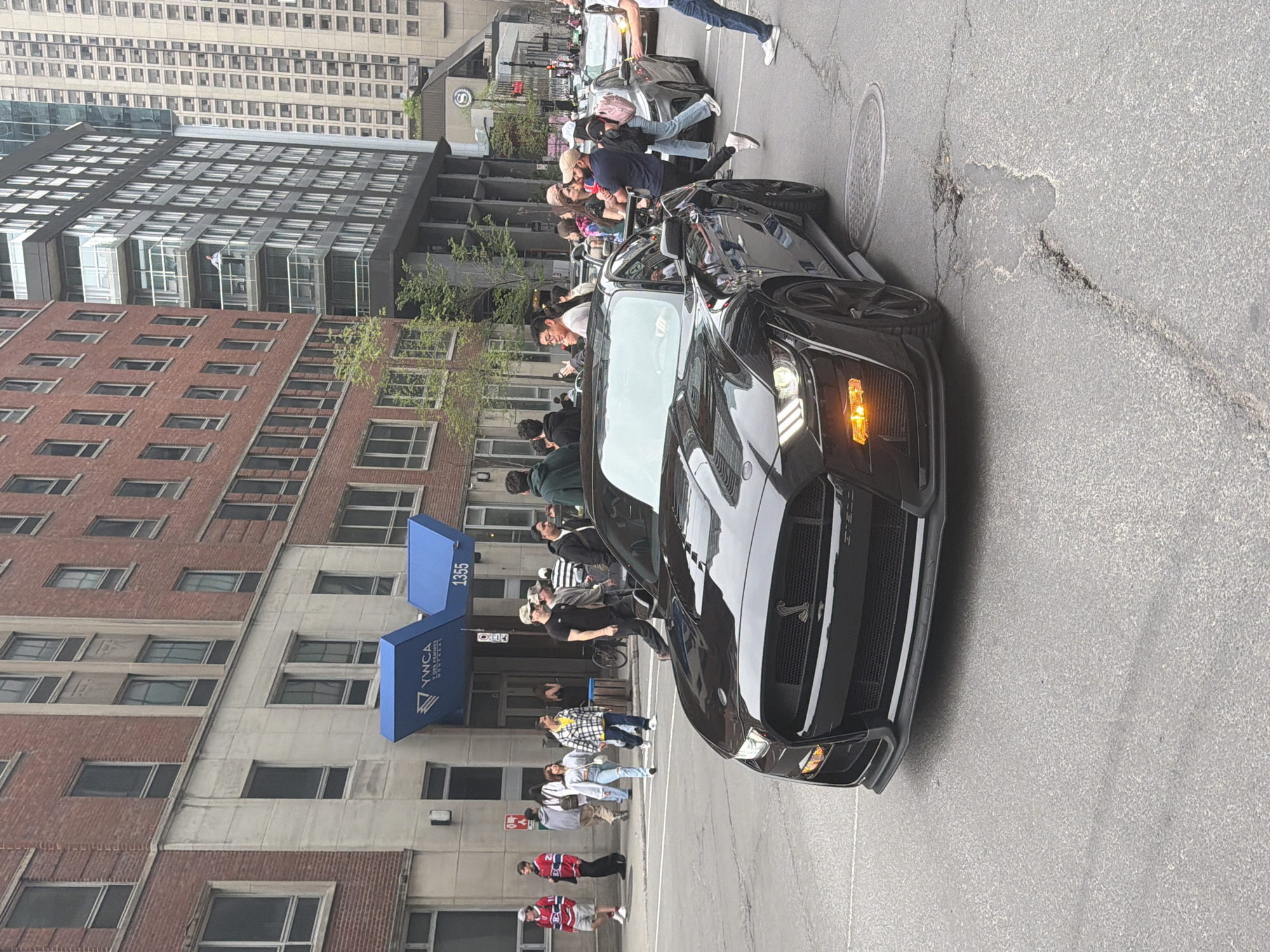 The image shows a black Ford Mustang with a crowd of people gathered around it in Montreal, Canada. The car is parked in the street in front of a building with a blue awning that reads "YWCA Montreal" and the address "1355." There are several pedestrians on the street, some looking at the car and some walking past. In the background, there are tall buildings and trees. The weather appears to be overcast. There is a manhole cover in the street, and cracks in the asphalt. The Sherato Hotel is visible in the background.