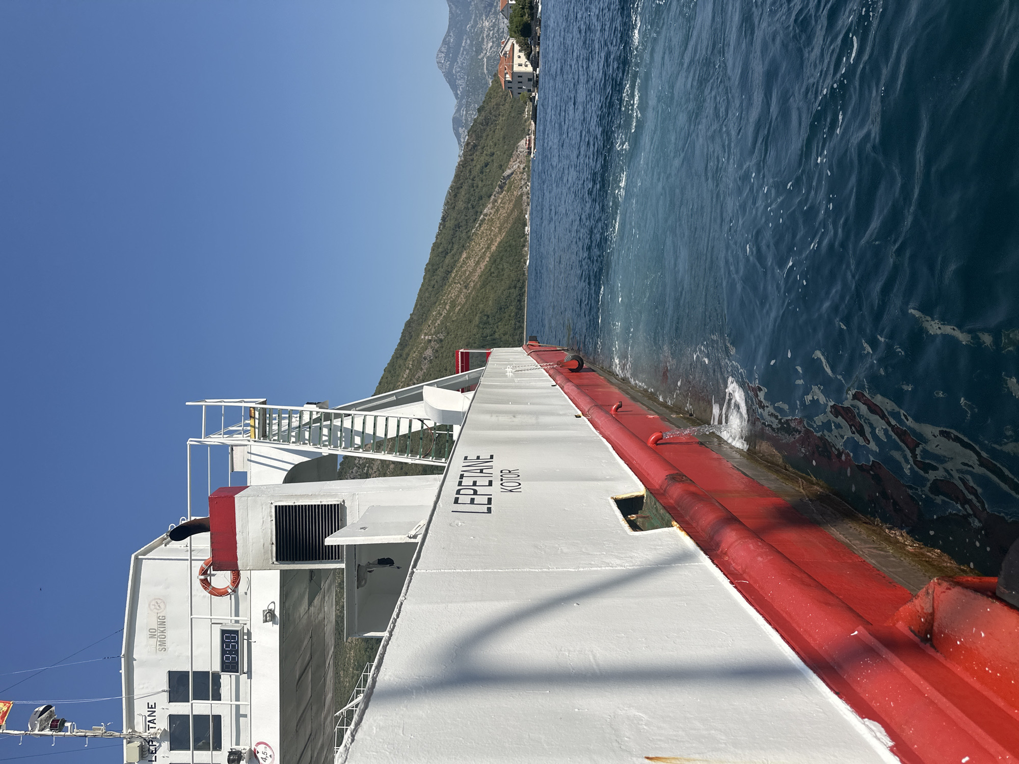 A side view of the ferryboat "LEPETANE KOTOR" is shown as it moves through the Bay of Kotor near Lepetane, Montenegro. The ferry, painted white with a red hull, is in motion, creating a wake in the blue water. A digital clock on the ferry displays "9:50". A "NO SMOKING" sign is visible on the upper deck. The surrounding environment includes a green, tree-covered hillside and mountains in the background, along with a few buildings on the shore. The sky is clear and blue.