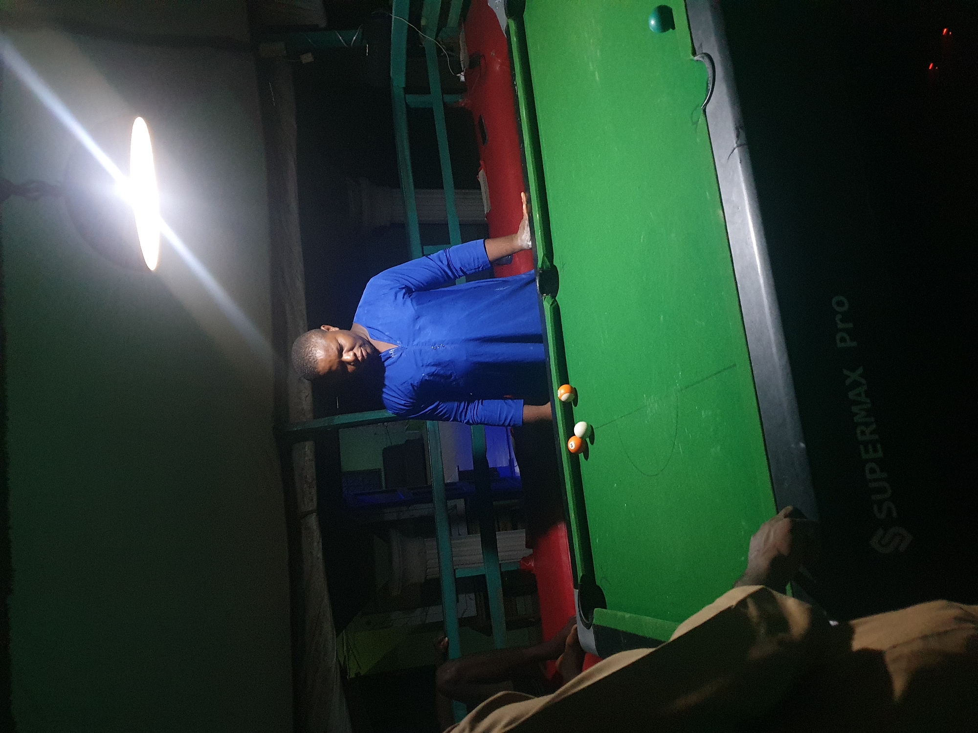 In Lokoja, Nigeria, a man in a blue traditional outfit is standing behind a green billiard table, looking down at the balls. Three billiard balls are on the table: one white, one orange, and one white with an orange stripe. Another person, wearing a brown garment, is partially visible on the left side of the frame, seemingly playing pool. The scene is illuminated by a bright overhead light, casting shadows and highlighting the details of the billiard table, which has "SUPERMAX Pro" inscribed on its side. The environment appears to be an indoor or covered outdoor space, with a dimly lit background suggesting a casual setting.