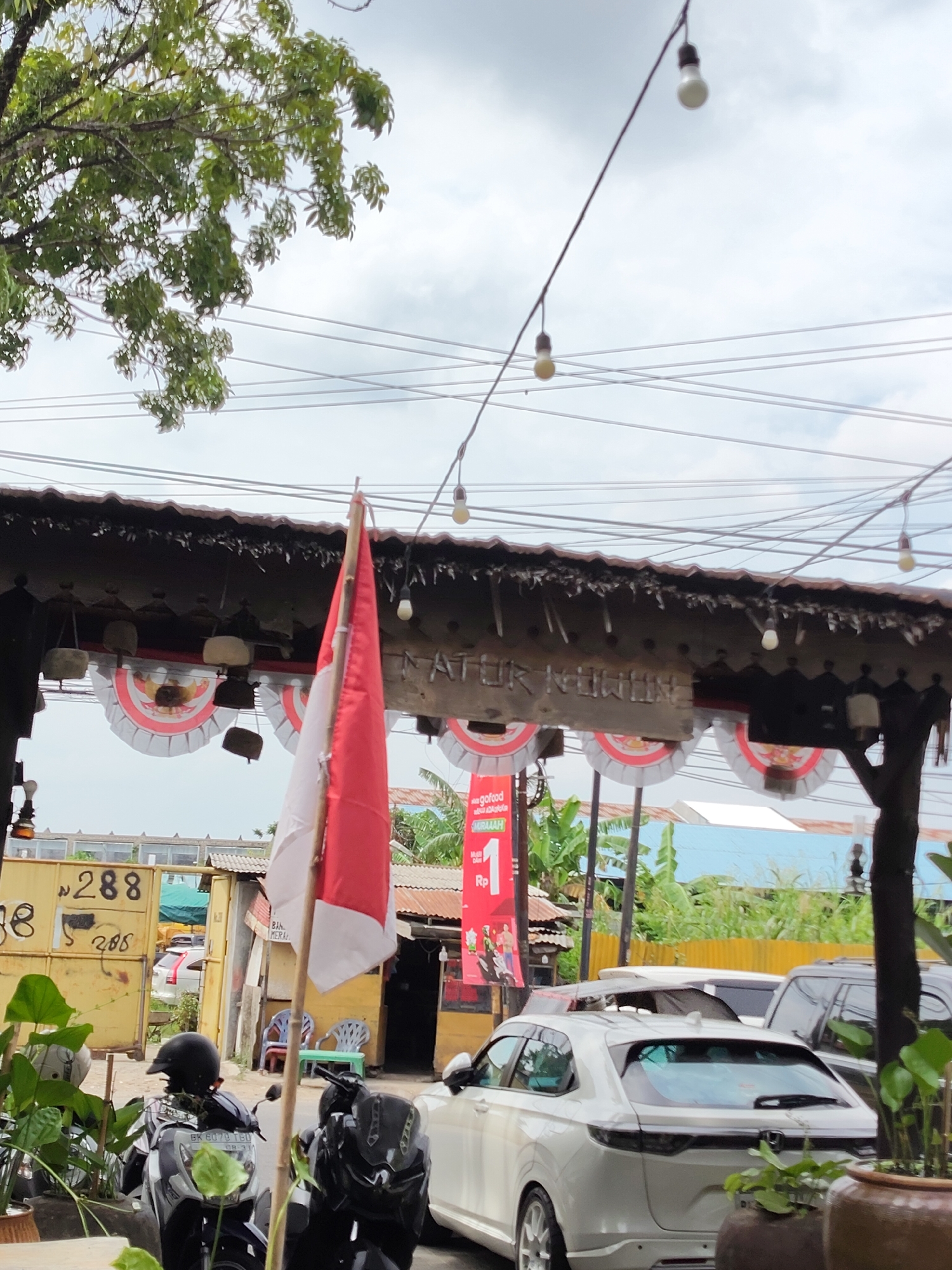 The image depicts an outdoor scene in the city of Medan, Indonesia, likely on a slightly overcast day. In the foreground, two motorcycles are parked, one black and one predominantly white and black. A red and white Indonesian flag is prominently displayed on a pole to the left of the motorcycles, partially obscuring a wooden sign overhead that reads "NATUR NUHWIN". Decorative paper fans with red and white circular patterns are hung along the underside of the wooden structure.
Behind the motorcycles and flag, a yellow metal gate or container is visible with the number "288" and "288" inscribed on it. Beyond the gate, a white car is parked, and further in the background, there are buildings with corrugated metal roofs and lush green vegetation, including banana trees. Overhead, a tangle of electrical wires crisscrosses the sky, with several light bulbs strung along them. The overall impression is of a street-side establishment or marketplace in Medan.