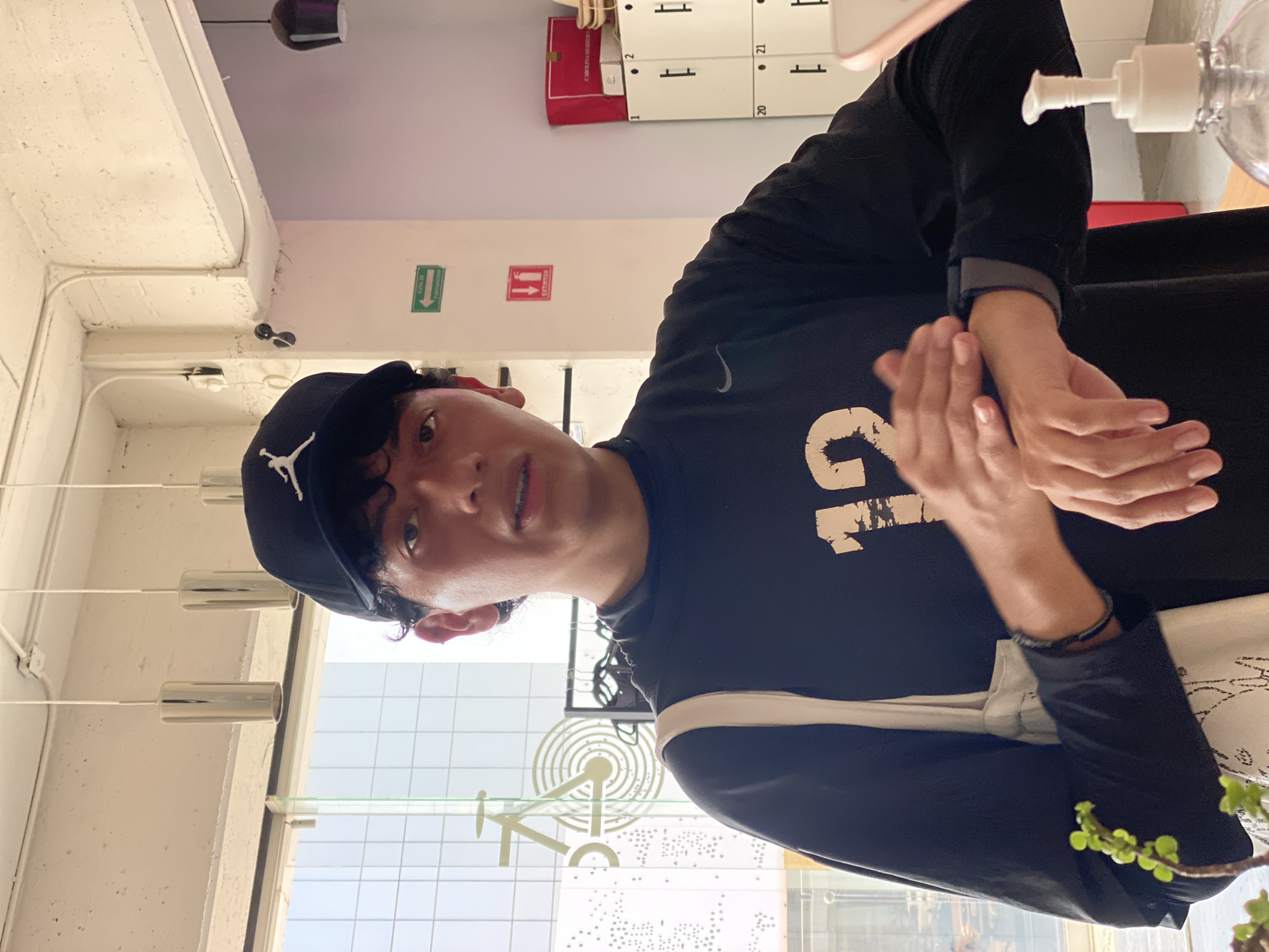 A young Hispanic male in a black Jordan baseball cap, a long-sleeved black shirt with a large off-white graphic and a white strap visible over his shoulder, is dispensing hand sanitizer. The background indicates a business interior with lockers, signage, and a view of a city street. A design of the city of Puebla is visible through the window, pinpointing the location as Puebla, Mexico. The individual is standing in front of a window with architectural features associated with Puebla, including a specific architectural representation of the city.