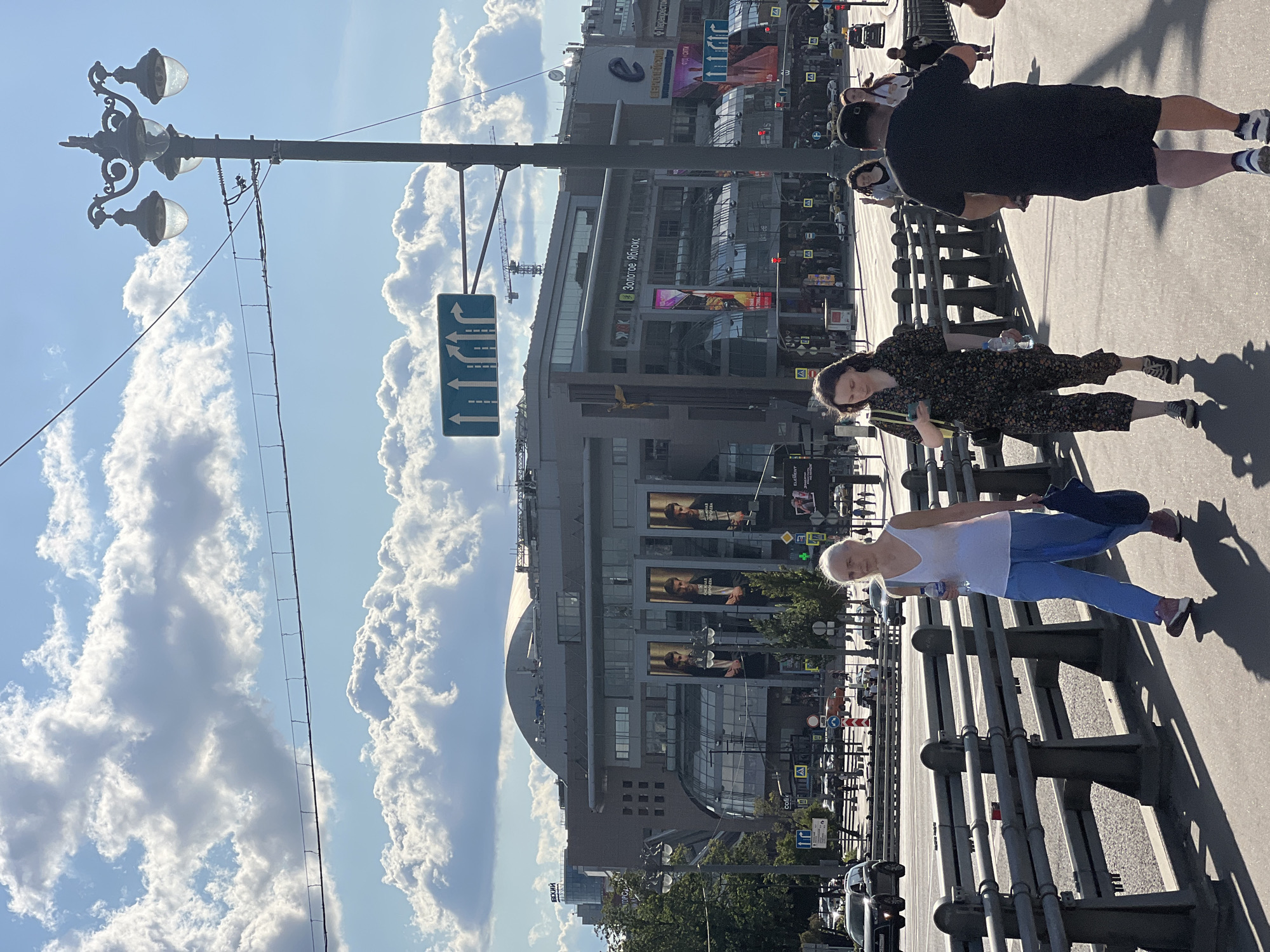 The image shows a sunny day in Moscow, Russia, on a bridge. A woman in a white tank top and blue pants walks on the right side of the frame, holding a water bottle and a dark bag. To her left, a woman in a dark floral print jumpsuit walks while looking at her phone. In the background, there is a large building with a curved roof, a street with cars and traffic lights, and a lamppost. Several people are visible walking on the bridge and crossing the street. The sky is blue with fluffy white clouds.