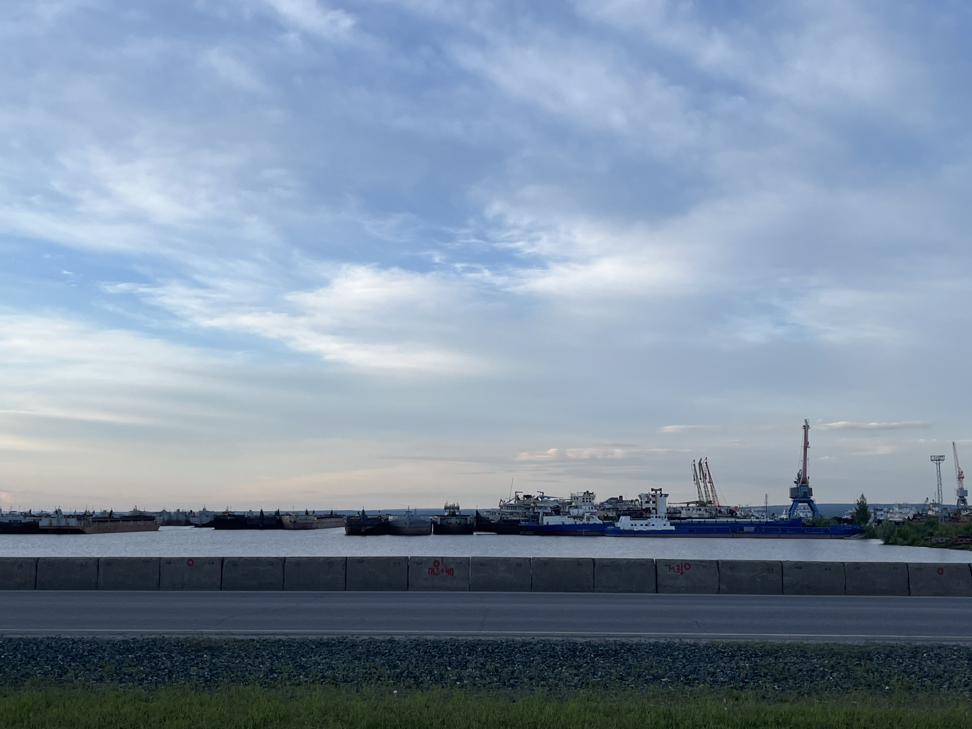 The image shows a harbor scene in Chaikovsky, Russia. In the foreground, there is a gray concrete barrier and a road. Beyond the barrier, there is a body of water with several barges and ships docked. In the background, there are industrial cranes and buildings. The sky is overcast with clouds.