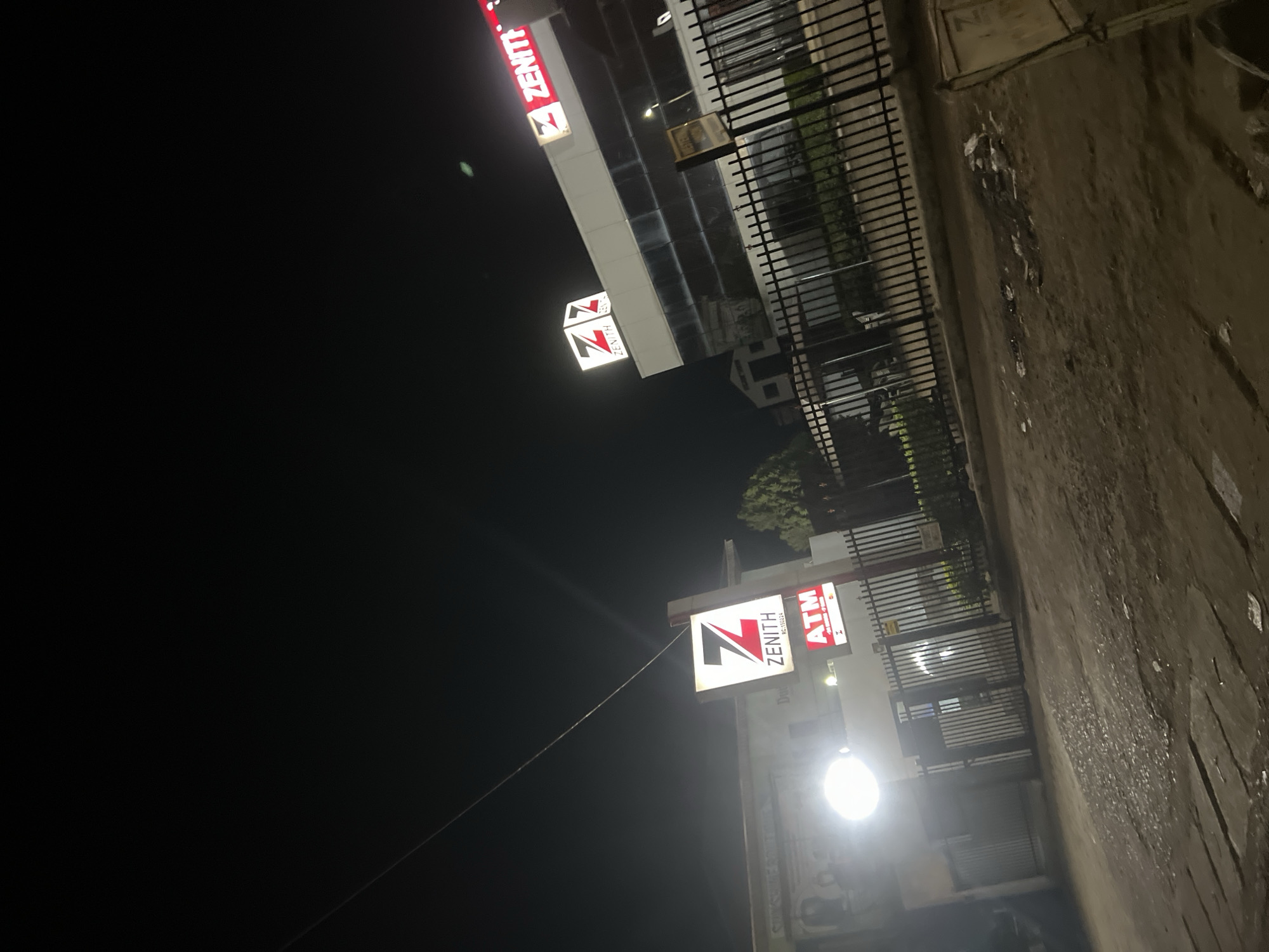 The image presents a nighttime street-level view in Jos, Nigeria, featuring two commercial buildings identified as "Zenith Bank" branches.
On the left, a building with a light-colored facade is illuminated by a prominent, bright circular white light fixture. Below this light, a rectangular sign displays the "Zenith" logo in red and white, with the text "RC:150224." Directly beneath it is a smaller red sign indicating "ATM," accompanied by Mastercard and Visa logos. A black metal gate, partially open, secures the entrance to this property.
To the right, a larger, multi-story building exhibits a modern architectural style with large glass windows and white paneling. Multiple illuminated "Zenith" signs, featuring the red 'Z' logo and text, are mounted on its roofline and facade. A street light is visible in the middle ground, slightly to the right of the central axis. A black metal fence runs along the front of this building, separating it from the unpaved foreground. Behind the fence, some greenery is discernible. A brown sign, partially obscured but possibly reading "Western Union," is positioned near the fence line in front of the right building.
The foreground consists of an unpaved, uneven dirt surface with visible patches of debris and discolored areas. A thin, diagonal line, likely a power or utility cable, is visible in the upper left portion of the dark sky. No individuals, vehicles, or specific actions are depicted in the scene.