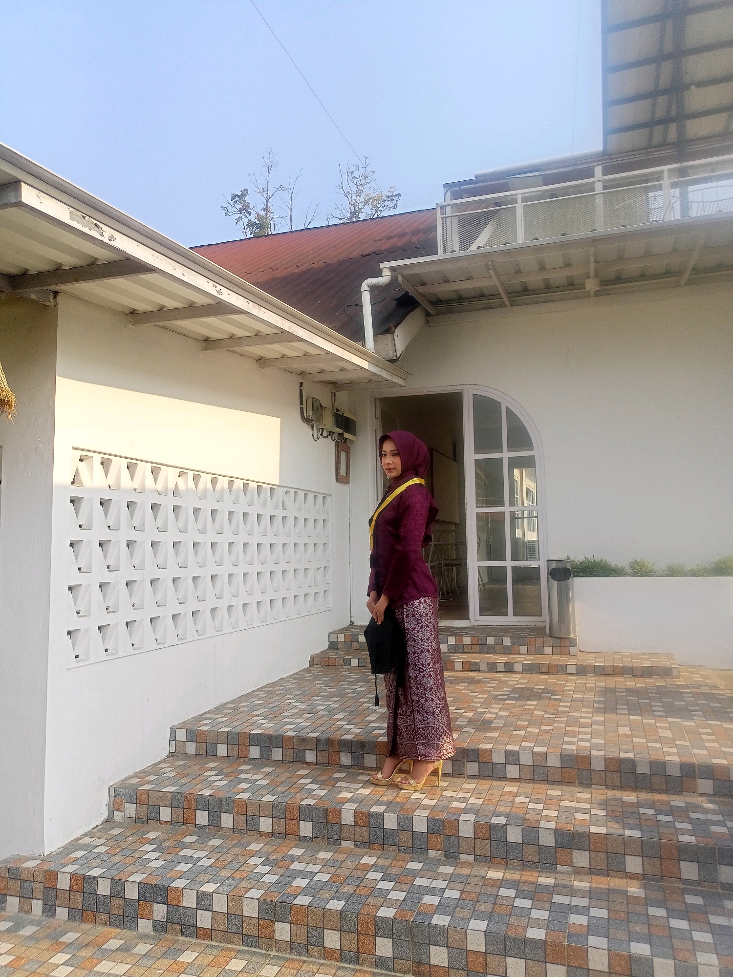 A young woman, dressed in a maroon brocade kebaya and a matching hijab with a yellow sash draped over her shoulder, stands on a tiled staircase outside a white building. She holds a black graduation cap in her left hand and a black clutch bag. The building features white latticework on its wall, a white-framed arched door with glass panes, and a white railing on an upper level. The staircase is covered in square tiles of various earth tones. The scene is bathed in bright, natural sunlight, casting shadows. The background shows a clear blue sky and some bare tree branches. This scene appears to be located in a residential or commercial area in Blambangan, Indonesia.