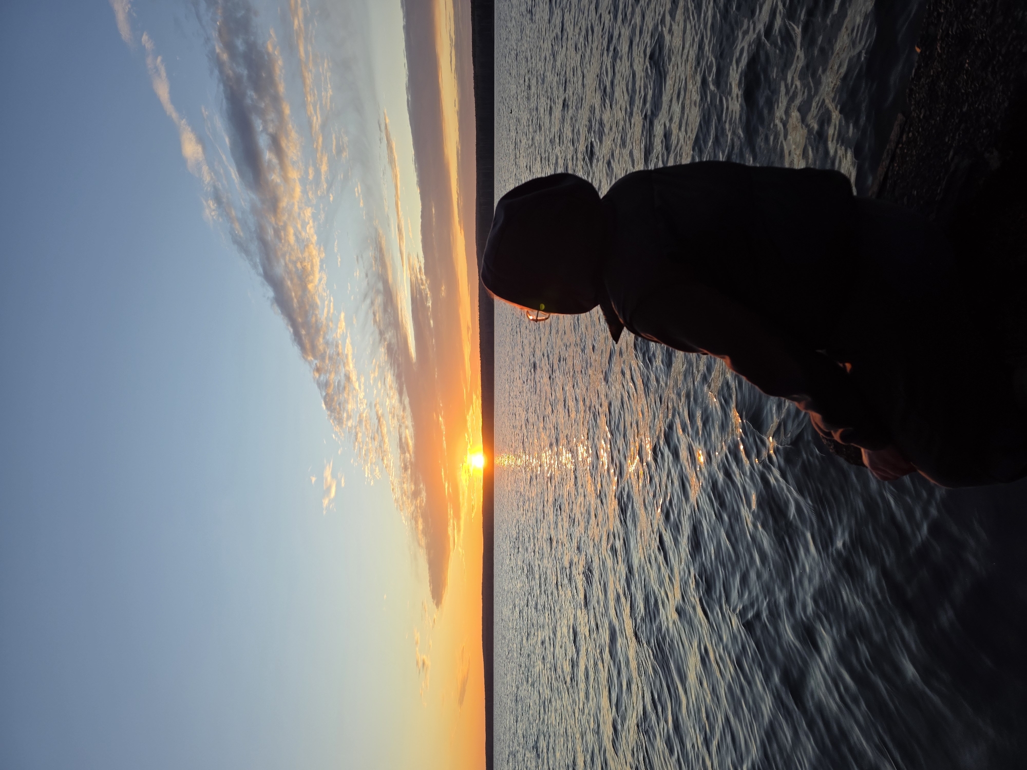 The image captures a sunset scene with a person silhouetted against the water. The location is likely Chaikovsky, Russia, given the context. To the left is a colorful sky with wispy clouds, transitioning from blue to orange. The sun is setting directly on the horizon, casting a bright reflection on the water. To the right of the horizon is a body of water, with light reflecting off of its surface creating a shimmering effect. In the foreground, on the right, is a dark silhouette of a person wearing a hooded jacket, facing the water. The person appears to be standing near a rocky or textured edge.