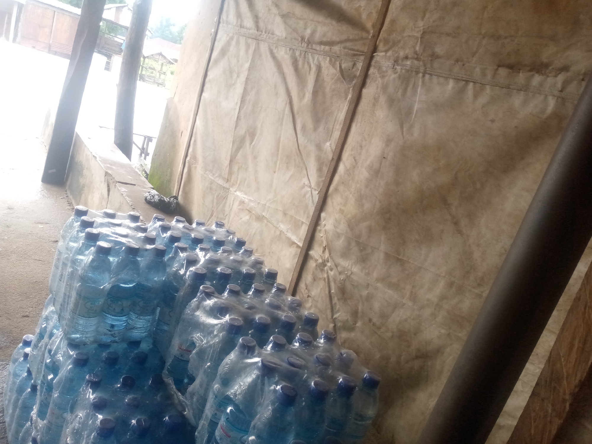 The image shows a large stack of bottled water, wrapped in plastic, stored in an open, somewhat dilapidated structure. The structure appears to be partially enclosed by a large, tan-colored tarp that is supported by wooden poles. The ground is a rough, concrete-like surface. In the background, there are glimpses of wooden buildings and vegetation, suggesting a rural or semi-rural environment within or near the city of Serti, Nigeria. The lighting is somewhat dim and overcast, possibly indicating an indoor or shaded area. There are no people visible in the image. The overall impression is one of storage and possibly a distribution point for the bottled water.