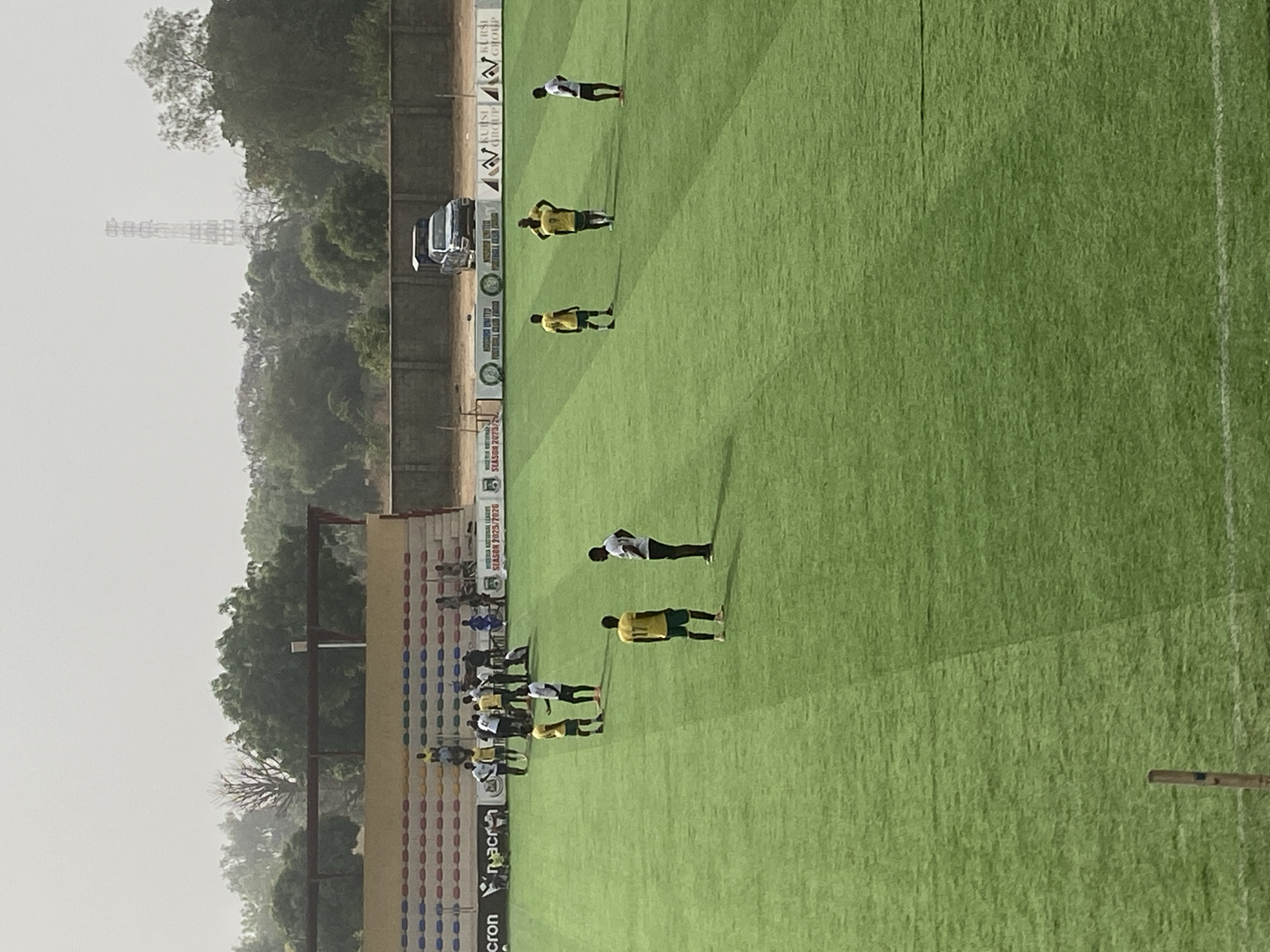 The image shows a soccer field in Tudun Wada, Nigeria. Multiple players in yellow and black uniforms are standing on the field, with some wearing white uniforms. A group of people are in the stands. A white van is parked behind a perimeter wall with advertisements. A tall transmission tower is visible in the background.