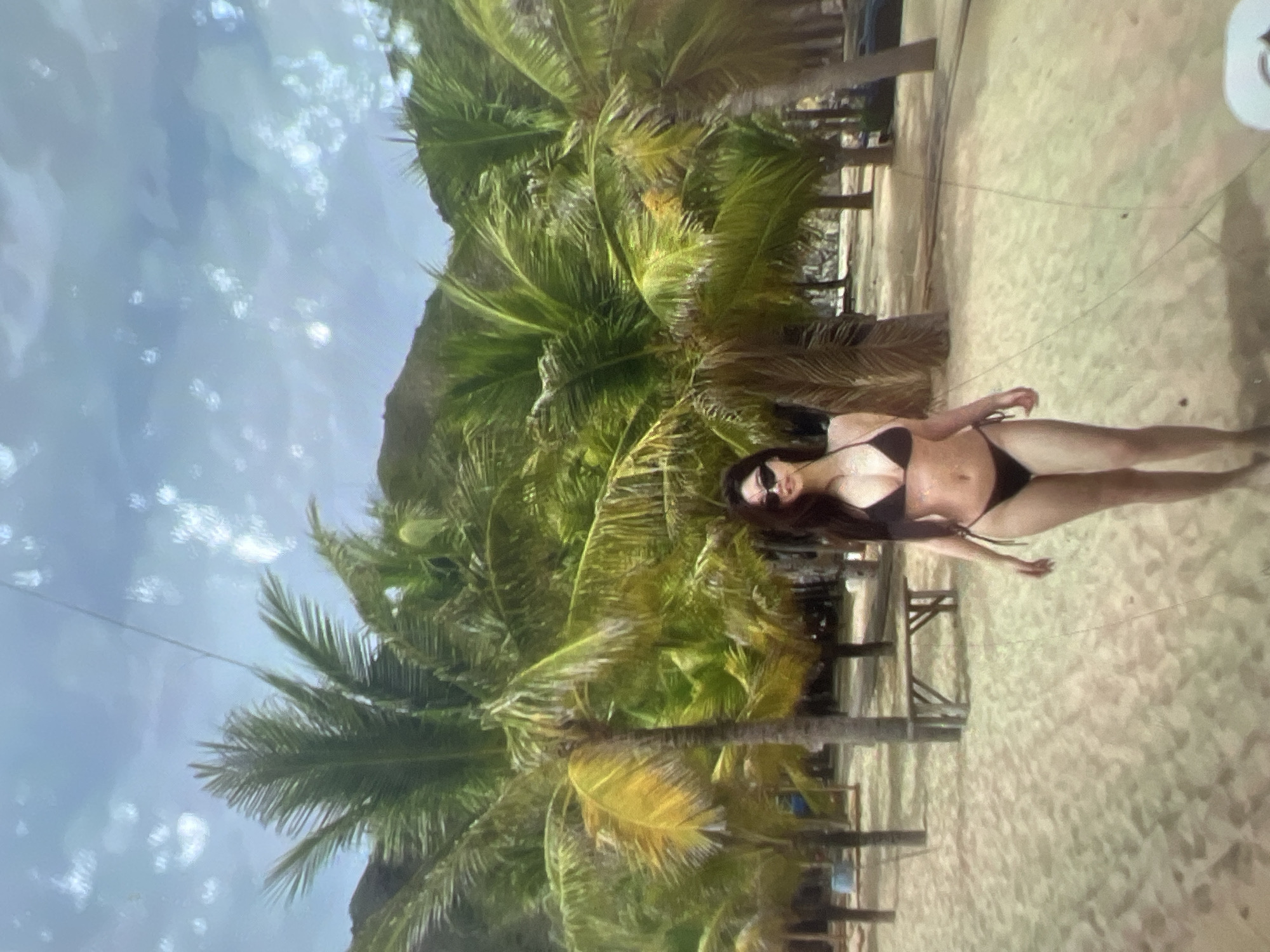A young woman wearing a black bikini and sunglasses stands on a sandy beach. She is facing the camera with her arms at her sides. Behind her, there are numerous palm trees and a light blue sky with white clouds. A wooden bench is visible in the background. The location is identified as Kara-Dıykan, Kyrgyzstan, which is an unusual setting for palm trees and beaches, suggesting it might be a man-made environment or a misidentification.