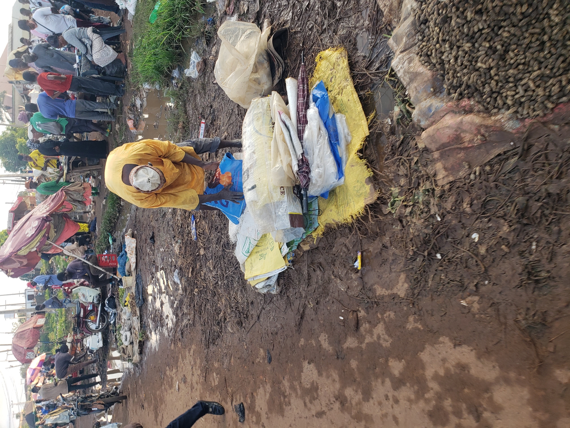 This is a medium-angle, ground-level, eye-level shot of an outdoor market scene in Kakuri Gwari, Nigeria. The ground is muddy and littered with debris. In the foreground, a man wearing a yellow shirt and a white cap is bent over, sorting through a pile of plastic bags and what appear to be umbrellas. He is surrounded by a collection of bundled items, some wrapped in yellow and white plastic. To the right, a large pile of unshelled peanuts is visible, resting on a reddish, textured surface. In the middle ground, several people are standing and interacting, some near motorbikes and market stalls. One person is sitting on a red crate. In the background, more people, market umbrellas, and buildings can be seen. The overall impression is one of a bustling but somewhat disorganized outdoor marketplace.