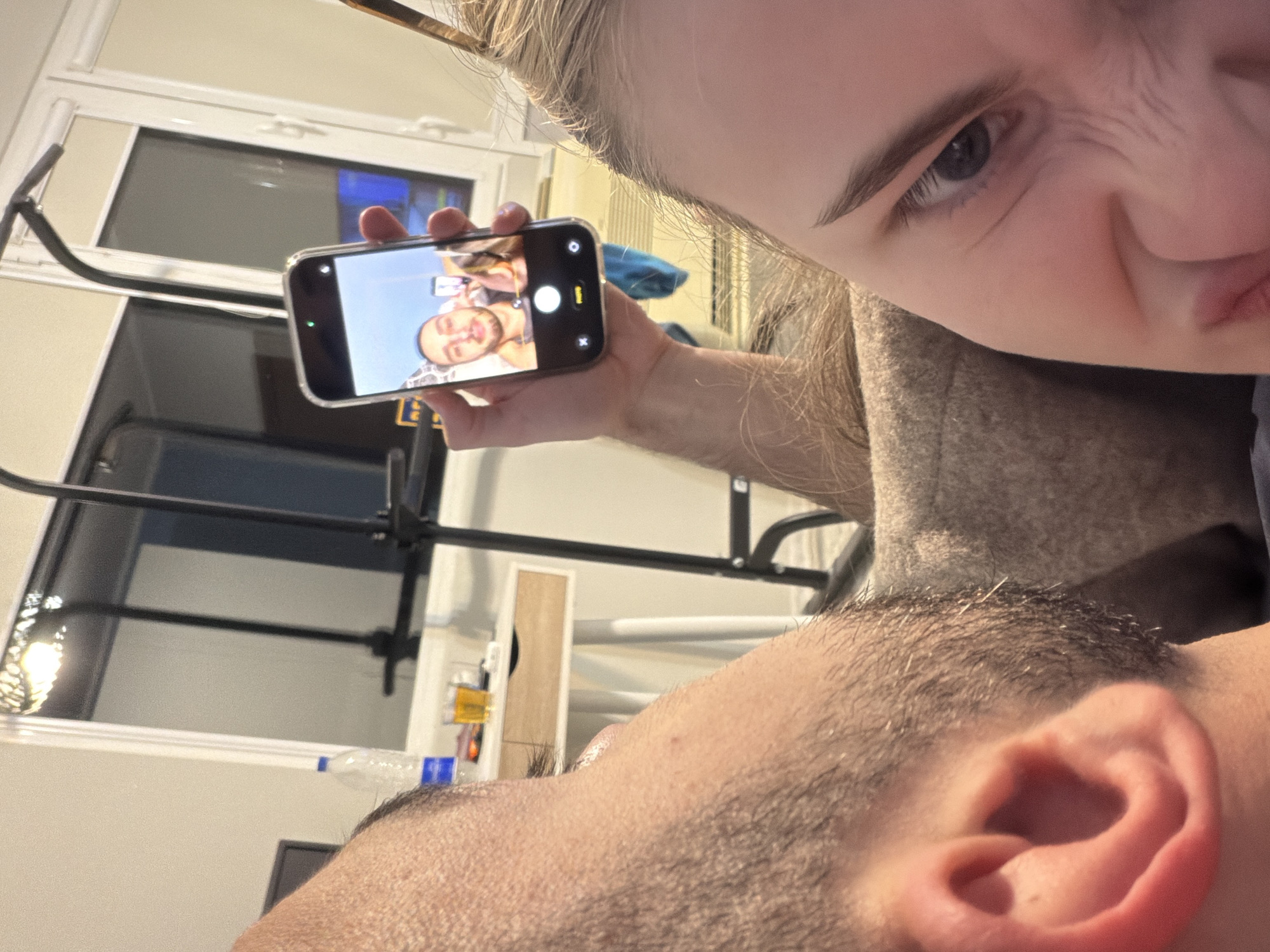 The image captures an indoor scene in Plovdiv, Bulgaria, showing two individuals in close proximity as one takes a smartphone selfie.
In the left foreground, the upper left profile of a male subject is visible, encompassing his right ear, side of his head, and neck. He exhibits short, dark hair and light stubble along his jawline. His skin tone is fair.
In the right foreground, a female subject is partially visible, displaying her right eye (blue in color), eyebrow, forehead, and a portion of her scrunched lower face, creating wrinkles around her nose and mouth. She has light-colored (blonde or light brown) hair. She wears a light brown, textured garment. Her right arm is extended, holding a dark-colored smartphone enclosed in a clear protective case. The phone's screen displays a live camera preview, capturing the upper torso and face of a man with a light beard (likely the male subject) and a partial view of another person (likely the female subject) in the background. The phone interface indicates "PHOTO" mode.
The background reveals a light-colored desk or table positioned behind the male subject. On this surface, a plastic water bottle with a blue label, a clear glass containing an amber liquid, and a small, unidentifiable orange object are present. A light-colored drawer is integrated into the desk. Behind the desk, a large window with white frames and horizontal blinds is visible, reflecting a partially obscured, illuminated light fixture at the top left. A black metal structure, resembling exercise equipment (possibly a pull-up bar), stands near the window. Below the window on the right, a white horizontal unit, potentially an air conditioner or heater, is installed. The room features light-colored walls and appears to be illuminated by artificial light.