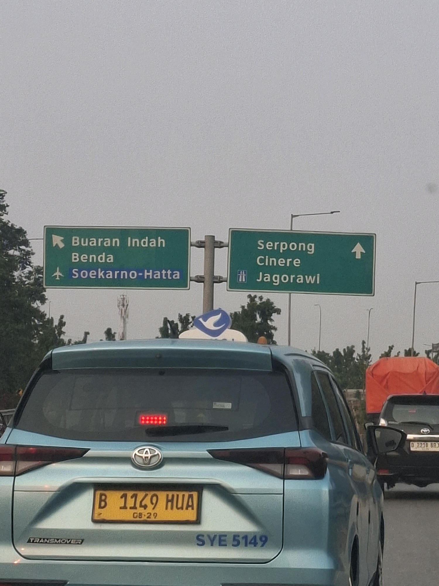 This is a medium shot of a road scene in Tangerang, Indonesia, captured from the perspective of being inside a vehicle. The foreground is dominated by the rear of a light blue SUV. The license plate of this SUV reads "B 1149 HUA" with "08-29" below it. Text on the rear of the SUV indicates "TRANSMOVER" on the left and "SYE 5149" on the right.
Above the SUV, a green overhead road sign is visible, split into two sections. The left section indicates "Buaran Indah Benda" with an arrow pointing left, and "Soekarno-Hatta" with an airplane icon pointing forward. The right section shows "Serpong Cinere" with an upward arrow, and "Jagorawi" with a highway icon. Trees and utility poles are scattered throughout the background.
In the middle ground, other vehicles are present, including a black SUV with license plate "D 2210 BRJ" and a truck covered with an orange tarp. The overall environment appears to be a highway or major road, with overcast skies contributing to diffused lighting.