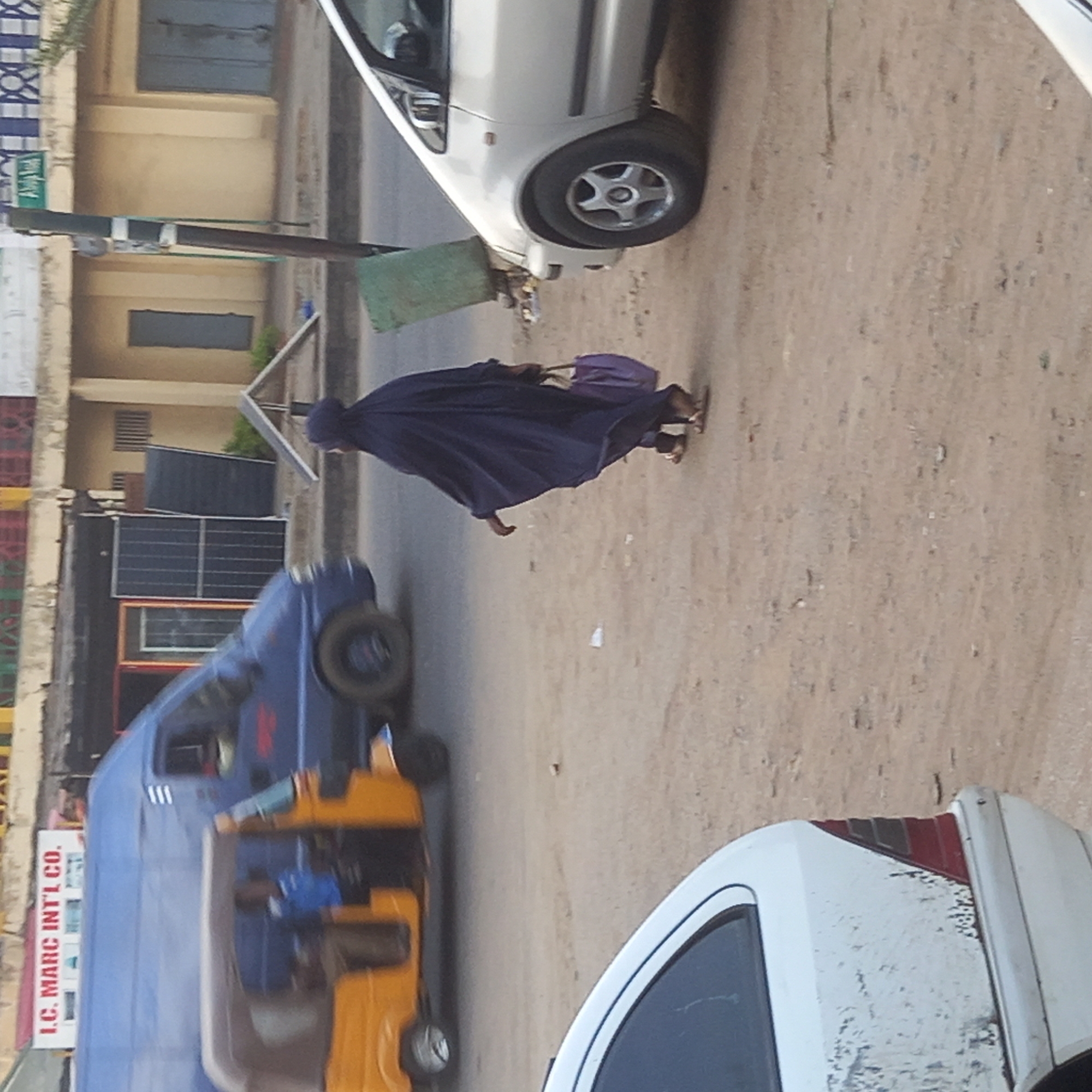 An individual wearing a long, dark garment walks along a street in Sardauna, Nigeria. A white car is parked on the right side of the frame, and another car is parked in the foreground. A blue and yellow tuk-tuk (three-wheeled vehicle) is also present in the scene. Buildings and street signs are visible in the background.