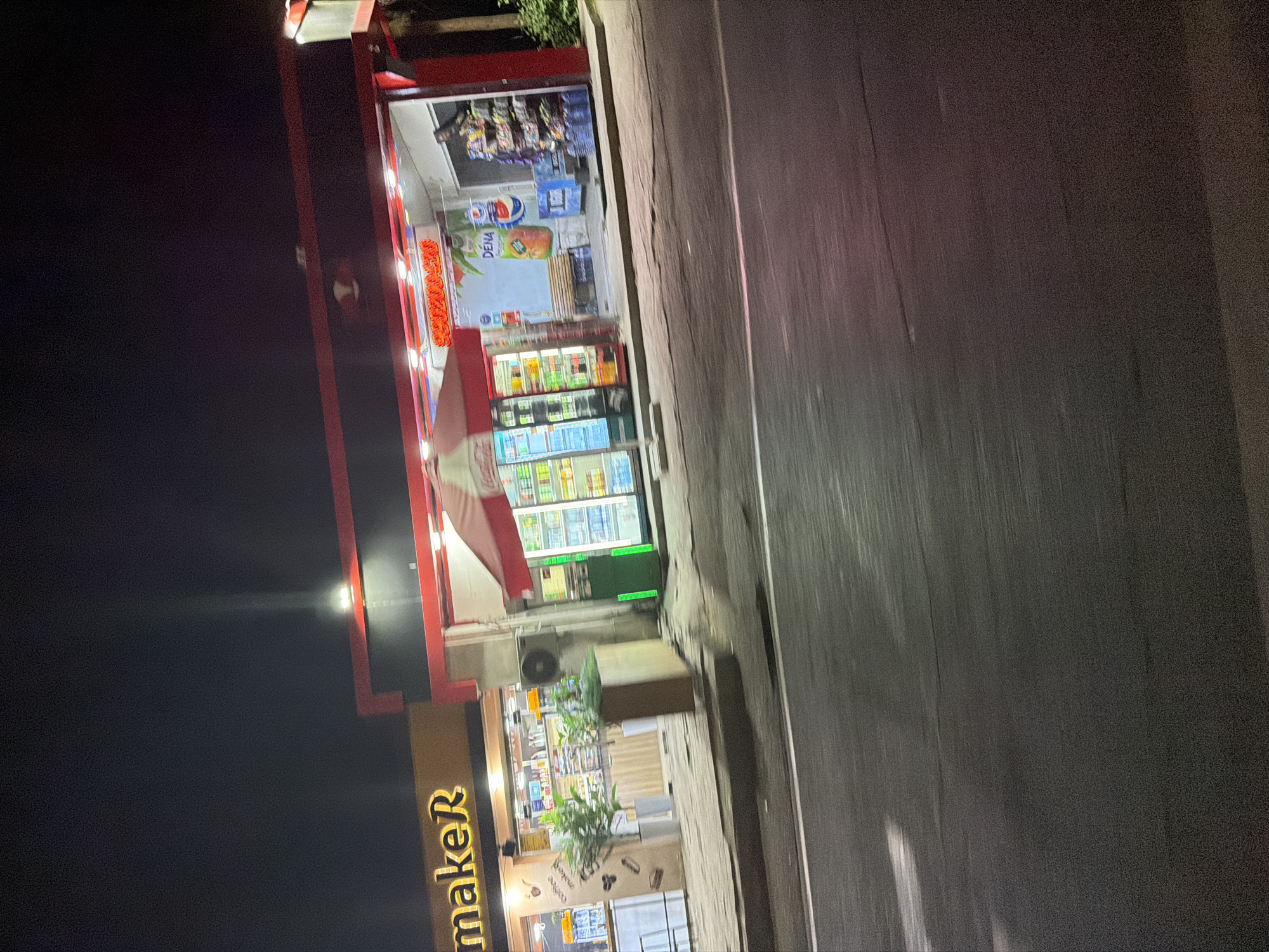 The image shows a nighttime street view of a convenience store in Tashkent, Uzbekistan. The store, named "makeR," features bright yellow lettering on its facade. To the right, a separate section of the store is brightly lit with red trim, displaying drinks and snacks. A red and white umbrella shades the front of this section. Inside, shelves are stocked with various products, and a Pepsi advertisement featuring the brand "DENA" is visible. A wooden bench is placed outside the store. The street in front of the store is paved and appears to be a road, with markings visible. The overall lighting suggests artificial illumination from the store and streetlights, contributing to the nighttime ambiance.