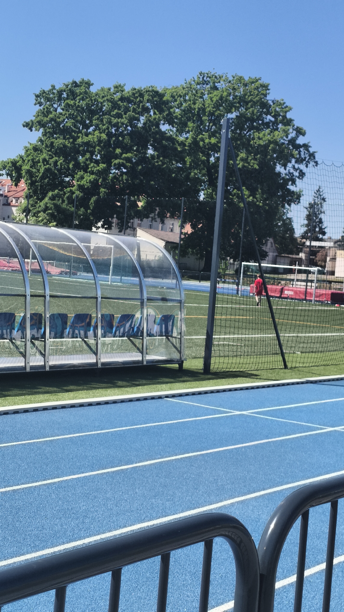 The image shows a view of a sports complex in Nowy Tomyśl, Poland, on a sunny day. In the foreground is a blue running track with white lane markings, partially obscured by a dark gray metal railing. Behind the running track is a soccer field with artificial turf. A clear plexiglass dugout with blue seating is visible on the left side of the field. A black net borders the field's edge. In the background, a person wearing a red shirt is seen walking on the soccer field, and a soccer goal is visible. Beyond the field and net, trees and buildings are apparent against a clear blue sky.