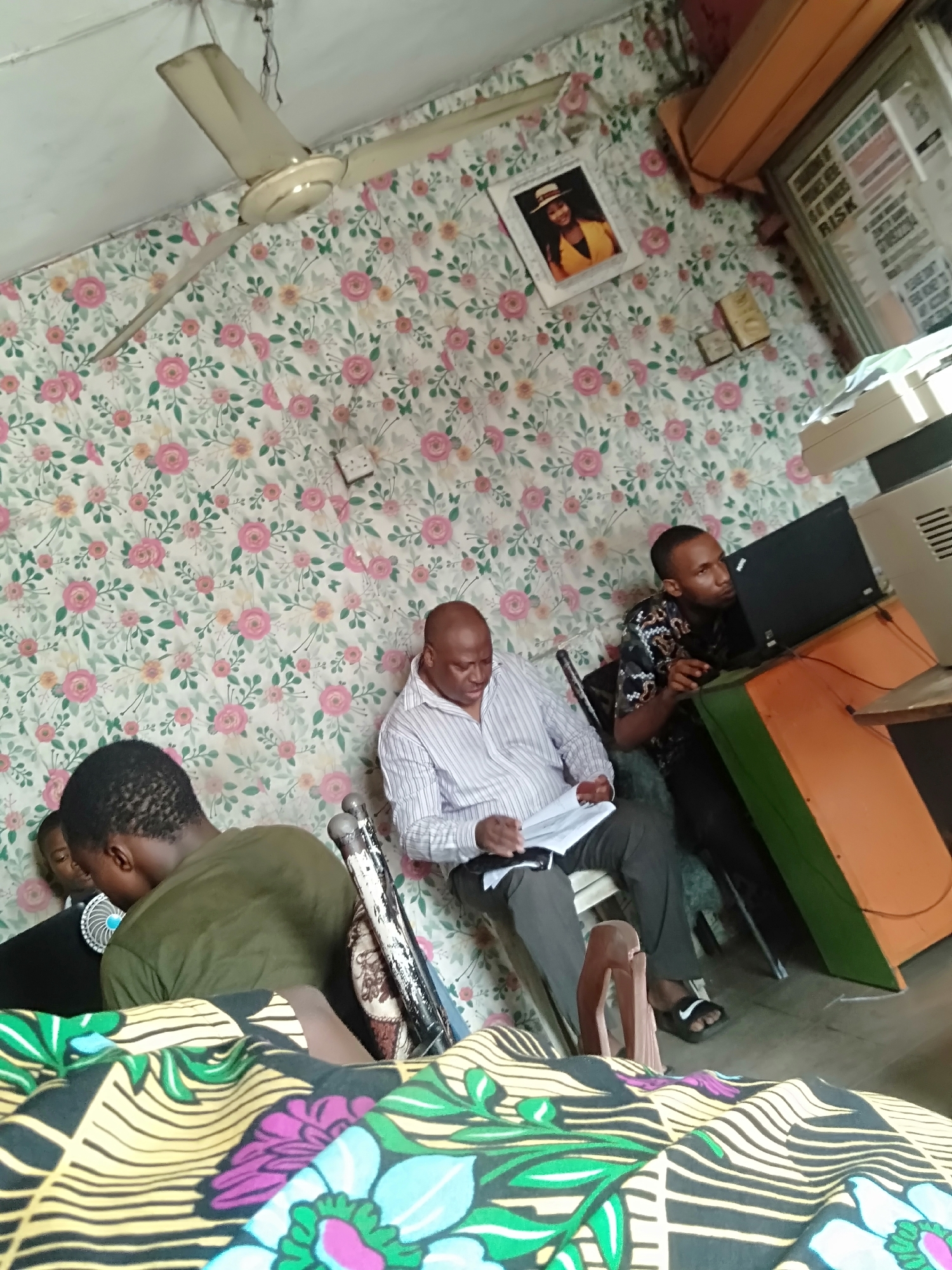 The image captures an indoor scene, likely a casual office or study environment, with a distinct tilt to the perspective. The dominant feature of the room is the patterned wallpaper, adorned with a busy floral design of pink, orange, and green roses and foliage on a light background.
Four individuals are present in the room. In the center, a bald, middle-aged man wearing a striped button-down shirt and dark trousers is seated, engrossed in reviewing papers he holds in his hands. He wears black slide sandals. To his right, a younger man with short dark hair, dressed in a patterned shirt, sits intently focused on a black laptop positioned on an orange and green desk. A light-colored printer or scanner is visible next to the laptop. In the left foreground, another person, seen from the back, wears a dark green t-shirt, partially obscured by a colorful patterned fabric that fills the bottom of the frame. Behind this person, a younger individual or child is partially visible, also facing away, with a small blue and white personal fan nearby.
The room features a white ceiling from which an old-fashioned, cream-colored ceiling fan hangs, with exposed wires near its mounting. On the wallpaper, above the bald man, a framed picture displays a woman in a yellow top and a straw hat. Below this picture, two small, possibly decorative, items are placed against the wall. A white electrical outlet is also visible on the patterned wall. On the far right, near the desk, some posters or papers are affixed to the wall, with fragments of text like "WHAT IS THE RISK," "WHAT," and "HOW" faintly discernible. The floor appears to be a plain concrete or tiled surface.
The overall impression is one of activity and work within a somewhat eclectic and functional setting in Shangisha, Nigeria. The lighting suggests it is daytime or a well-lit indoor space.