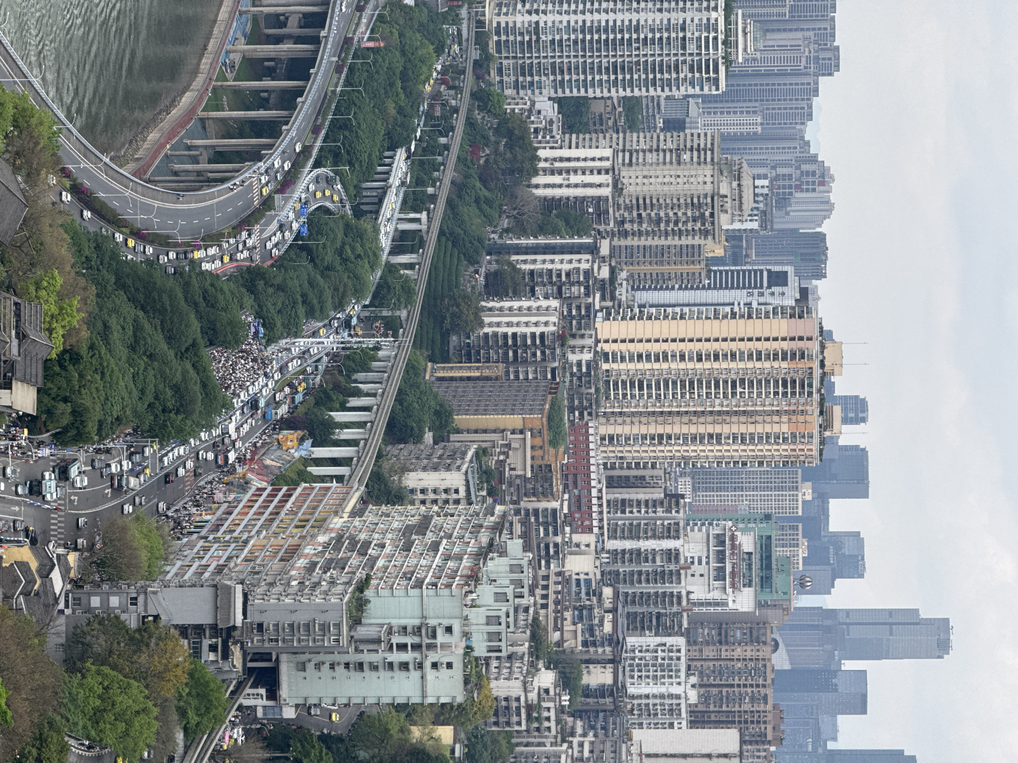 The image depicts an aerial view of Yuzhong District, China, rotated ninety degrees counterclockwise. Dense urban development is visible, including numerous high-rise buildings of varying heights and architectural styles. A multi-level highway curves along a river on the left. Vehicles, including cars and buses, are densely packed on the roadways, indicating heavy traffic. A monorail track runs parallel to the highway. Trees and vegetation are visible along the roads and in some areas between buildings. The sky is overcast.