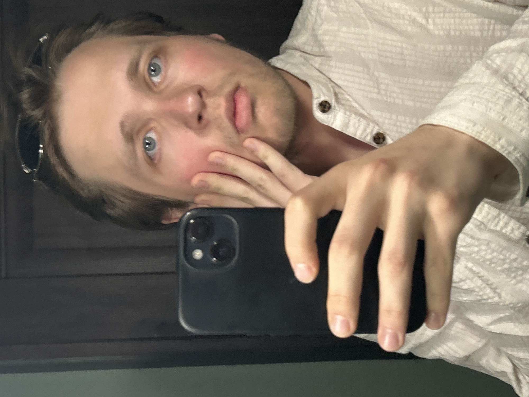 A selfie of a fair-skinned person with blue eyes, light brown hair, and stubble, wearing a white patterned button-down shirt. Sunglasses are perched on their head. The person is holding a black smartphone in their right hand, partially obscuring their face. Their left hand is touching their cheek. The background appears to be a dark, possibly wooden, surface, reflecting the image. The location is unknown.