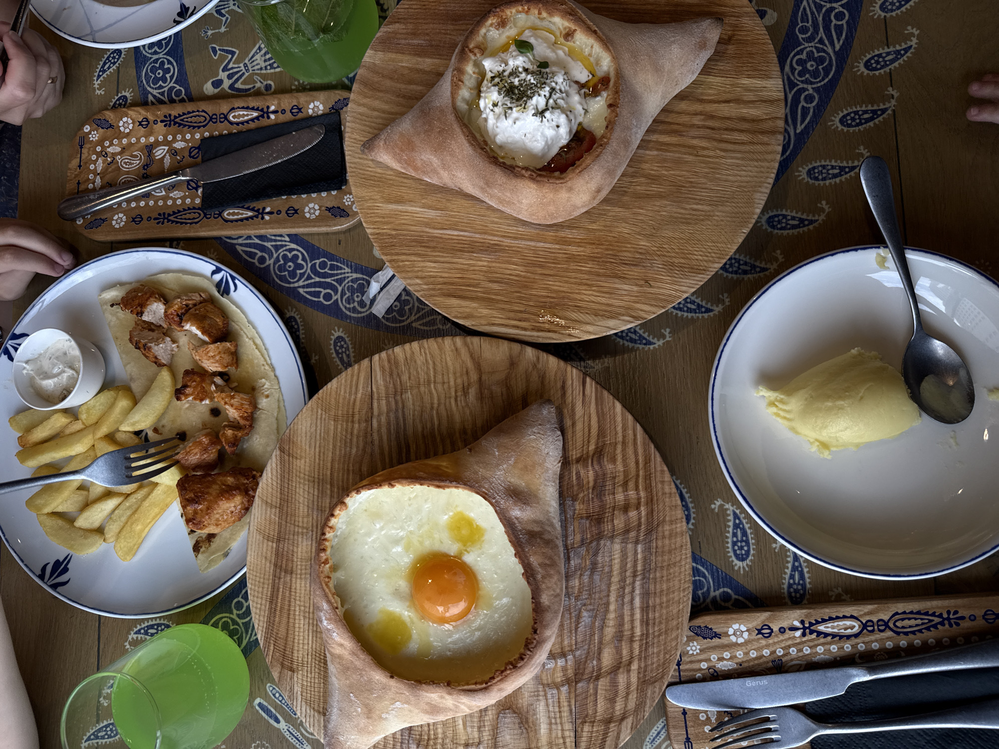The image displays a table setting for a meal, featuring several dishes and drinks. In the foreground, on the left, a fork and knife are placed on a black napkin. Adjacent to this, a white plate holds a dollop of yellow mashed potatoes and a spoon. To the right, a circular wooden board showcases a boat-shaped bread filled with melted cheese and topped with a fried egg. Another similar wooden board to the right holds another boat-shaped bread filled with cheese and white creamy topping, sprinkled with herbs.
Further back, a white plate contains french fries, pieces of grilled chicken, and flatbread. A fork is resting on the fries. To the left and right, there are glasses filled with a green beverage. The table is covered with a patterned tablecloth featuring blue paisley and floral motifs. The scene suggests a meal being served at a restaurant, possibly in Krasnoyarsk, Russia, given the prompt's context. There are hints of human hands in the background, indicating people are present and likely consuming the food. The lighting is natural, suggesting daytime.