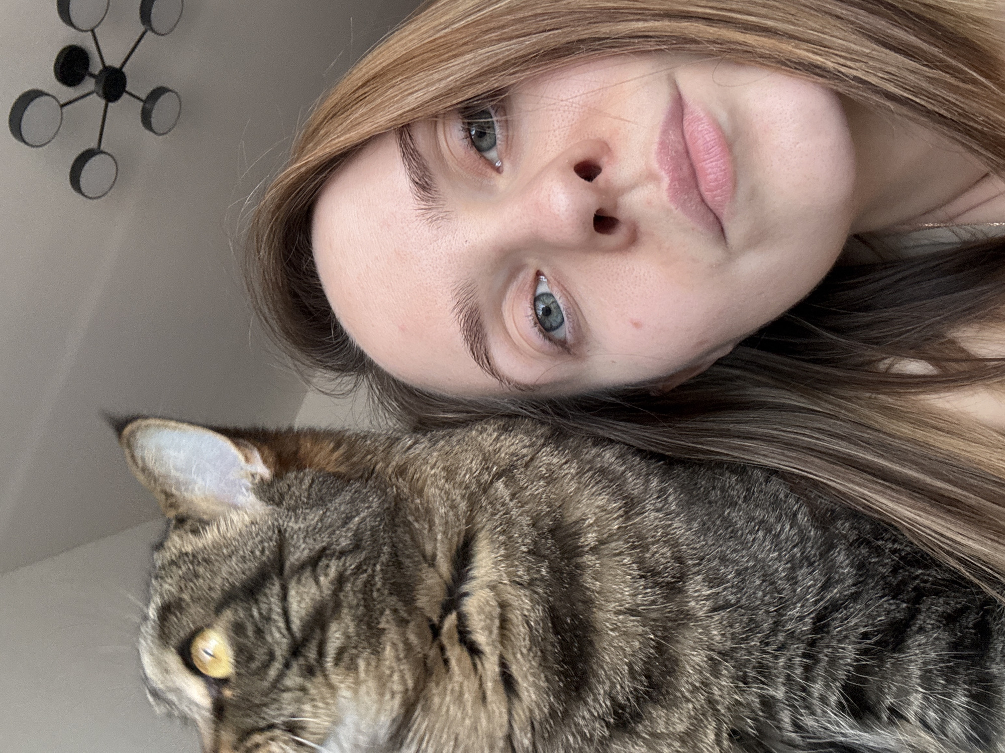 A close-up, high-angle interior shot captures a woman and a domestic tabby cat. The woman occupies the right half of the frame, with her light brown hair flowing downwards. Her pale complexion, light blue eyes, and visible pores are prominent. She looks directly at the camera with a neutral expression. A thin, light-colored necklace is visible below her chin. To her left, the tabby cat, distinguished by brown and black stripes and prominent yellow eyes, is positioned facing upwards and to the left of the frame. Only the cat's head, right ear, and upper torso are visible. The background consists of a plain light-colored ceiling. A modern black ceiling light fixture with five circular white or light-colored lamp covers, arranged in a sputnik style, is mounted in the upper right portion of the ceiling. The scene depicts a casual interaction between a person and a pet within what appears to be a residential environment. Specific visual identifiers to pinpoint a precise location within Perm, Russia, are not discernible.