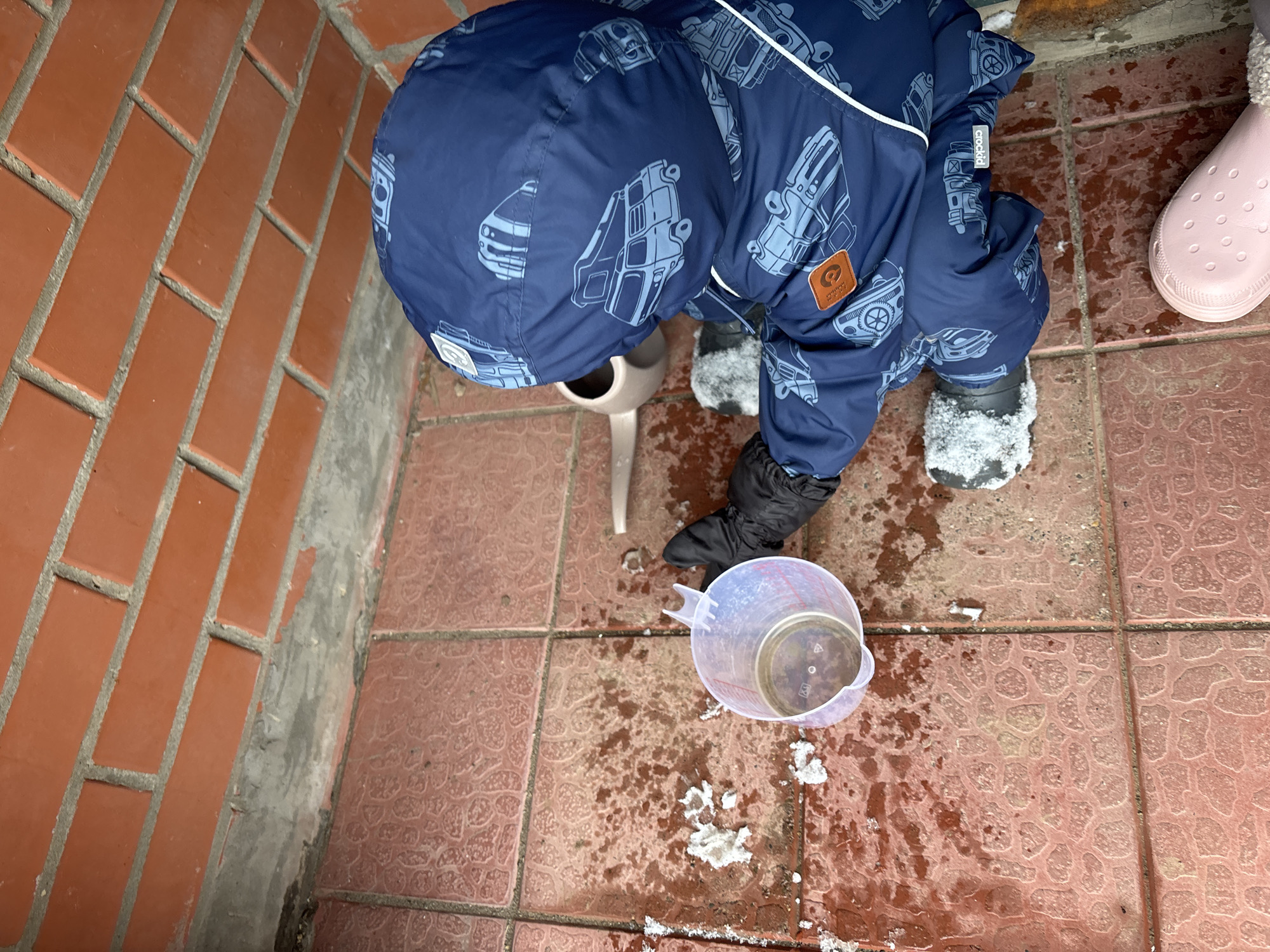 A child in a blue vehicle-patterned snowsuit and black boots stands on a tiled surface, next to a red brick wall. The tiled surface is wet and has patches of snow. The child is looking down at a clear plastic measuring cup filled with liquid. A light-colored watering can is nearby. A pink Croc-style shoe is visible at the edge of the frame. The location is identified as Ferma, Russia.