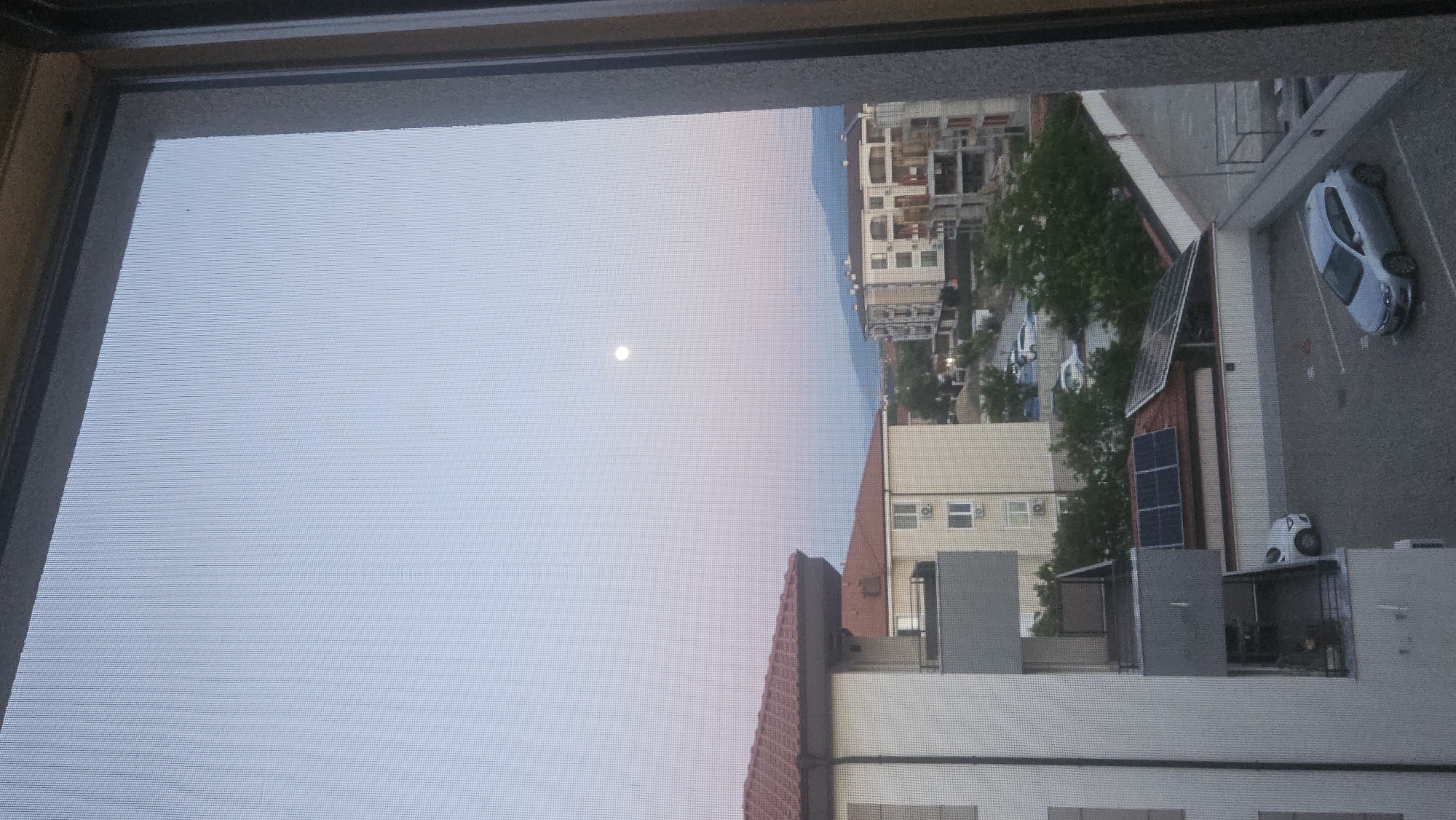 The image is a view through a window screen looking towards an urban setting. The sky is pale blue, and a full moon is visible. Below the horizon line of the sky, there are buildings and vegetation. The view is likely from a higher story, as the rooftops of buildings closer to the window are visible. There is a parking lot with cars at the right side of the image. Solar panels are visible on the rooftops of the buildings in the parking lot. Without further information, I cannot determine if this image is from Plovdiv, Bulgaria.