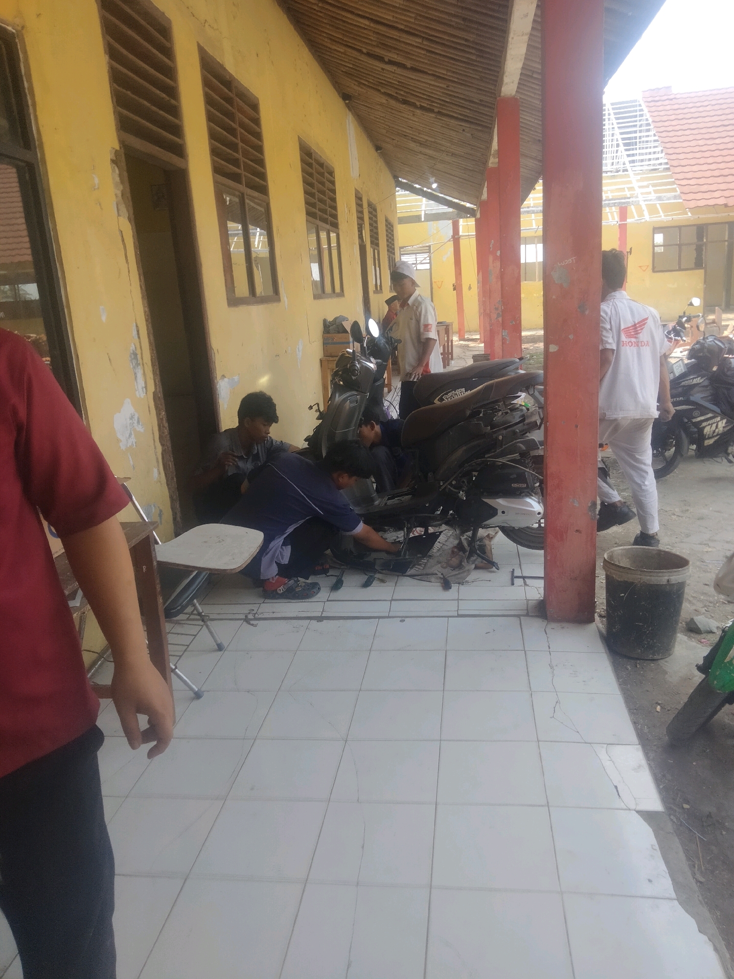 In Suka Sari, Indonesia, a group of individuals are gathered outside a yellow, weathered building with a bamboo-like roof structure. The building has several windows with wooden louvers and a dark-framed doorway. In the foreground, a person wearing a maroon shirt and dark pants stands on a tiled floor. The tiled floor shows cracks and discoloration. In the middle ground, several men are working on a dark-colored scooter parked on the ground. One man in a light-colored shirt and white cap is looking towards the scooter. Another man, wearing a dark blue shirt and black pants, is kneeling and appears to be working on the scooter. A third man, wearing a white shirt with a red "HONDA" logo on the back, stands next to the scooter and appears to be supervising or waiting. A black trash bin is located to the right of the scooter. Other scooters are visible in the background, and the area appears to be an outdoor space adjacent to the building.