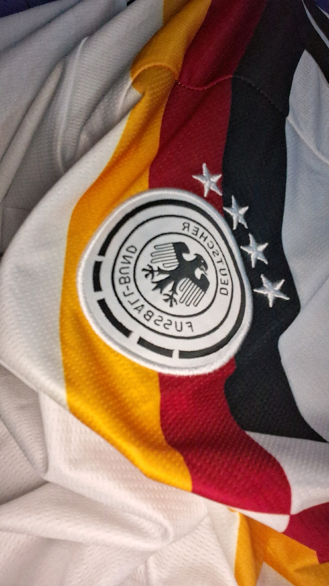 The image shows a close-up view of a German national football team jersey. The jersey features the colors black, red, and yellow, arranged in vertical stripes. A circular crest with a black eagle on a white background is visible on the left side of the jersey. The crest has text around it that appears to be in German. Three white stars are embroidered on the black stripe of the jersey, signifying past World Cup victories. The fabric of the jersey appears to be textured. The image does not provide any information about the city of Babakan, Indonesia, or any other environmental context.
