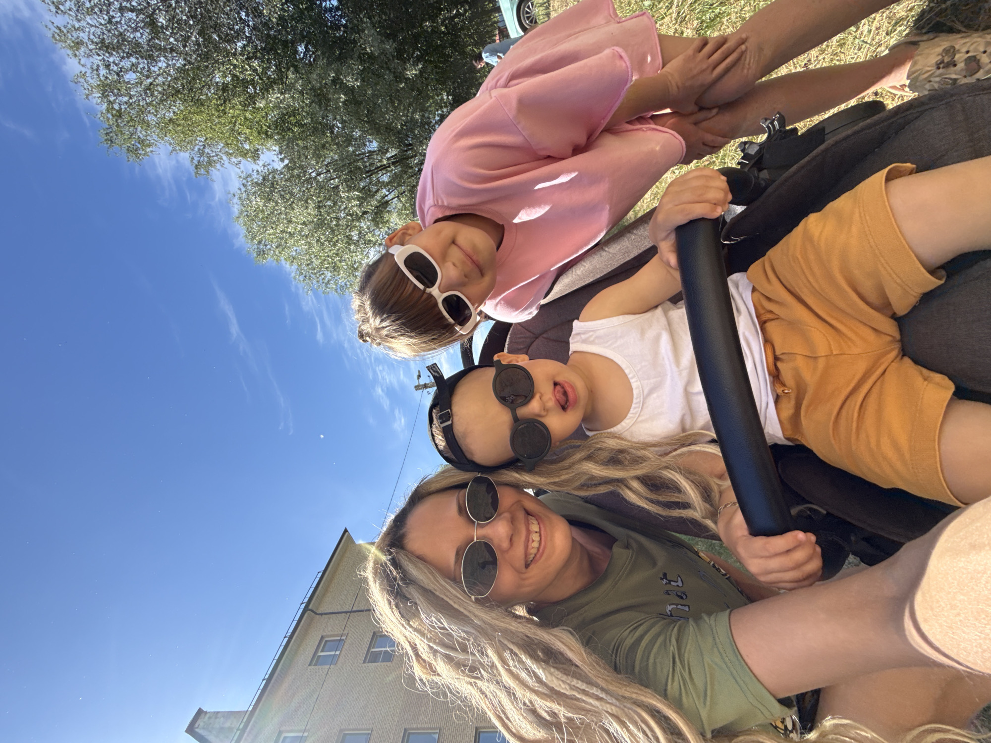 In Rusanovo, Russia, three individuals are captured in a selfie on a sunny day. A woman with long, blonde dreadlocks, wearing oval sunglasses and a green t-shirt, smiles at the camera. Next to her, a young child in a stroller wears round, black sunglasses and a white tank top with orange shorts, with their tongue sticking out. To the right, a girl with blonde hair pulled back in a bun sports white rectangular sunglasses and a pink oversized t-shirt. She leans in, her hand resting on her leg. The background features a brick building, lush green trees, and a clear blue sky with a few wispy clouds. The overall scene suggests a casual outdoor outing.