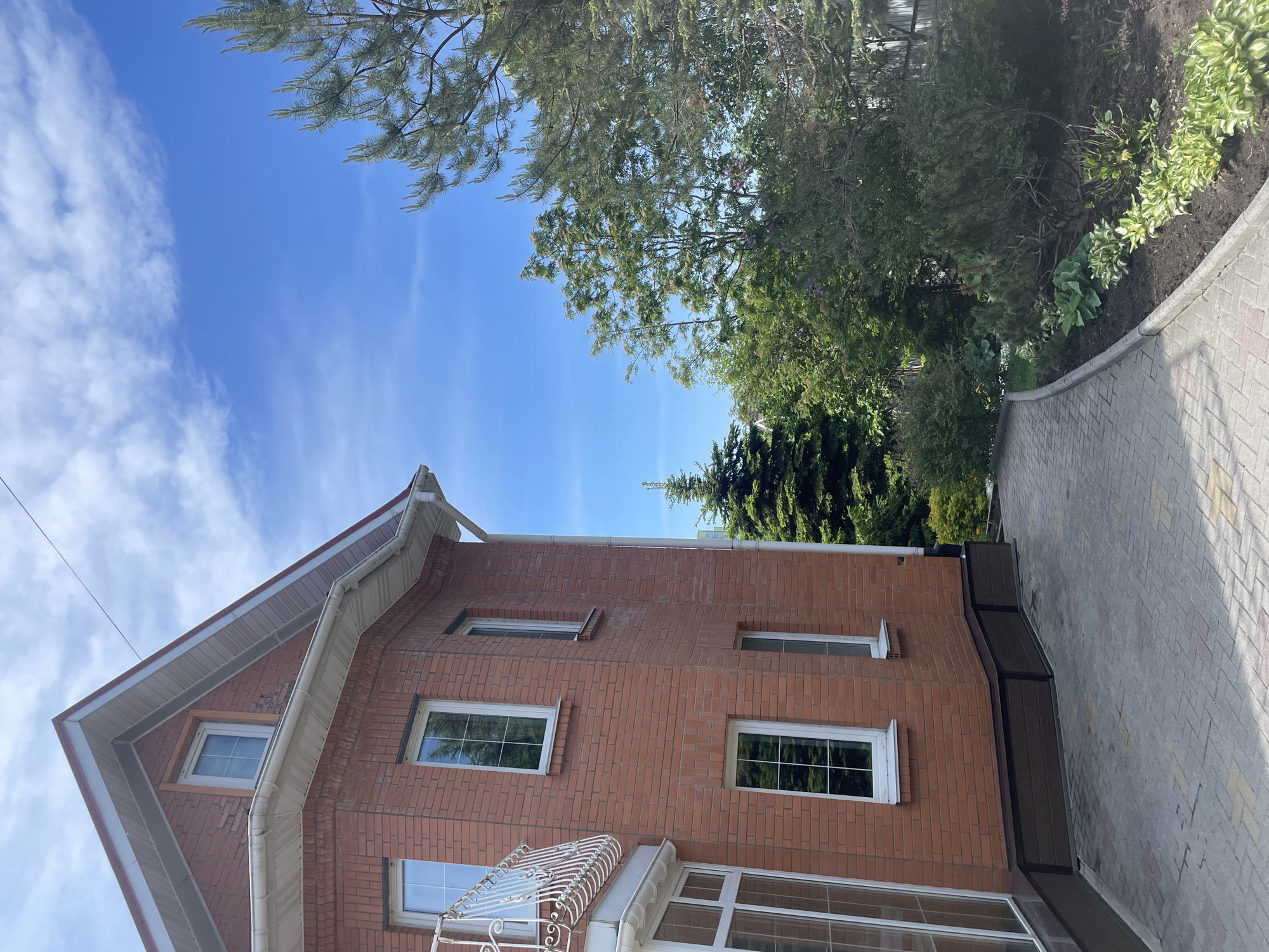 A two-story brick building is viewed from a low angle, seemingly from a street level. The structure exhibits red brick facade with white framed windows and a white-railed balcony. The sky is visible above the building, exhibiting a clear blue color with scattered clouds. Trees and shrubbery are present on the right side of the frame, growing alongside a paved sidewalk. The building's design includes a white trim along the roofline and a gable-style roof. The location is Vladivostok, Russia.