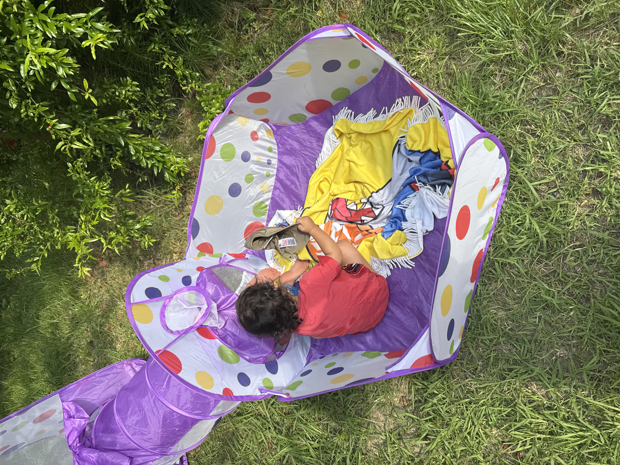 A young child with dark curly hair, wearing a red t-shirt and striped shorts, is lying inside a colorful pop-up play tent outdoors. The tent has a purple interior and is decorated with large polka dots in various colors, including red, yellow, blue, green, and purple. Several blankets and towels are piled inside the tent with the child. One blanket is bright yellow with a fringe, and another appears to have a cartoon character on it. The child is lying on their side, facing away from the camera, and seems to be interacting with something in their hands. The tent is set up on a grassy area, with lush green bushes and trees visible in the background. The sun is shining brightly, casting shadows and indicating it is likely daytime, possibly late morning or early afternoon. The overall scene suggests a playful and relaxed atmosphere, with the child enjoying playtime in a backyard or park setting. The location context of Sochi, Russia, places this scene within a region known for its warm climate and natural beauty. There is no visible text in the image.