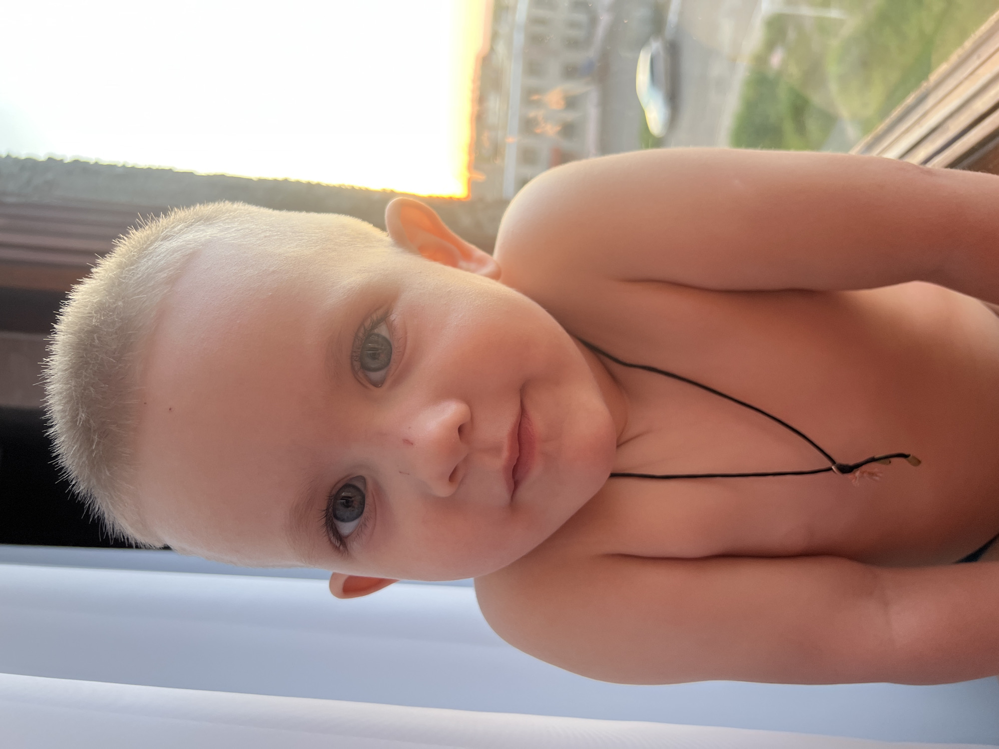 A close-up, slightly angled portrait captures a shirtless toddler with close-cropped blonde hair and gray eyes, peering directly at the camera. A simple black string necklace is visible around the child's neck. The background features a window offering a view of buildings, indicative of an urban setting, with an orange-tinged sky suggesting sunset. The reflections visible in the glass point to the location being Марковский, Russia. A vehicle is also visible in the distance through the window.