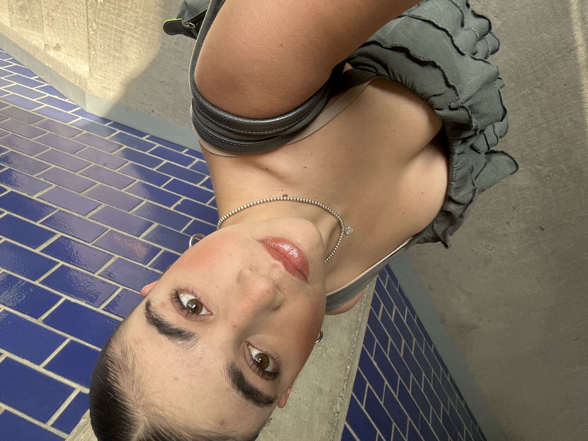 A close-up, upward-angled portrait captures a young adult female looking directly at the camera with a neutral expression. The image is rotated, presenting the subject in an inverted orientation. She has dark, pulled-back hair, prominent eyebrows, and brown eyes. Her face shows subtle makeup, including blush and glossy pink lipstick.
She wears a delicate beaded silver necklace with a small charm at its center, along with large hoop earrings. Her attire consists of a light-colored inner garment visible under a ruffled grey top or dress. A dark grey or black strap, possibly from a bag or another accessory, is draped around her upper arm.
The background is divided into two distinct sections. The lower part features a wall or floor covered in glossy, rectangular blue tiles arranged in a brick-like pattern. Above this, a rough-textured, light beige or grey concrete or stone surface is visible. The lighting is bright and even, suggesting either daylight or a well-lit indoor environment. No text is visible in the image. The scene is located in San Bernardino Tlaxcalancingo, Mexico.